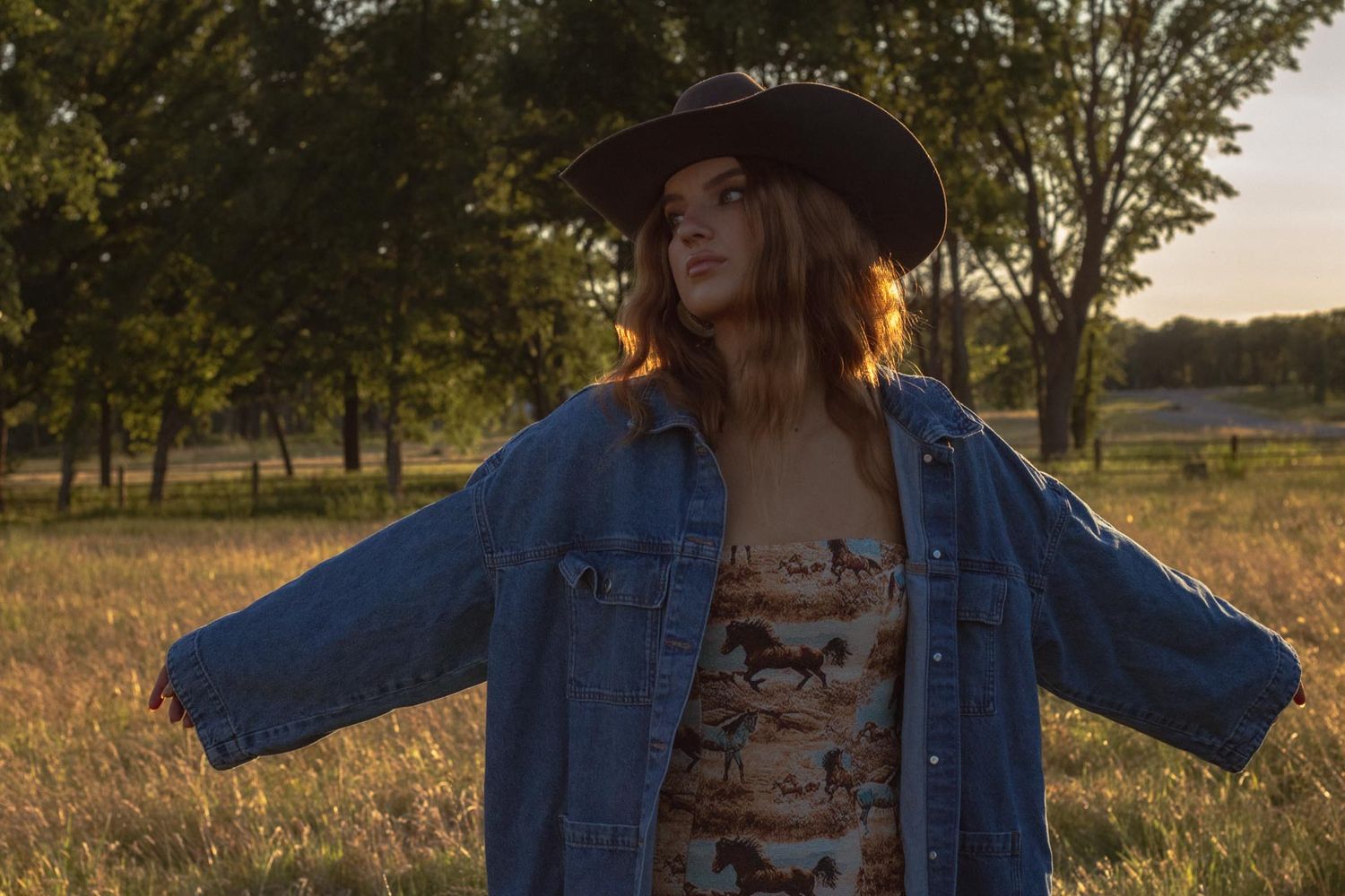 A person in denim and floral print clothing poses in a golden sunset field.