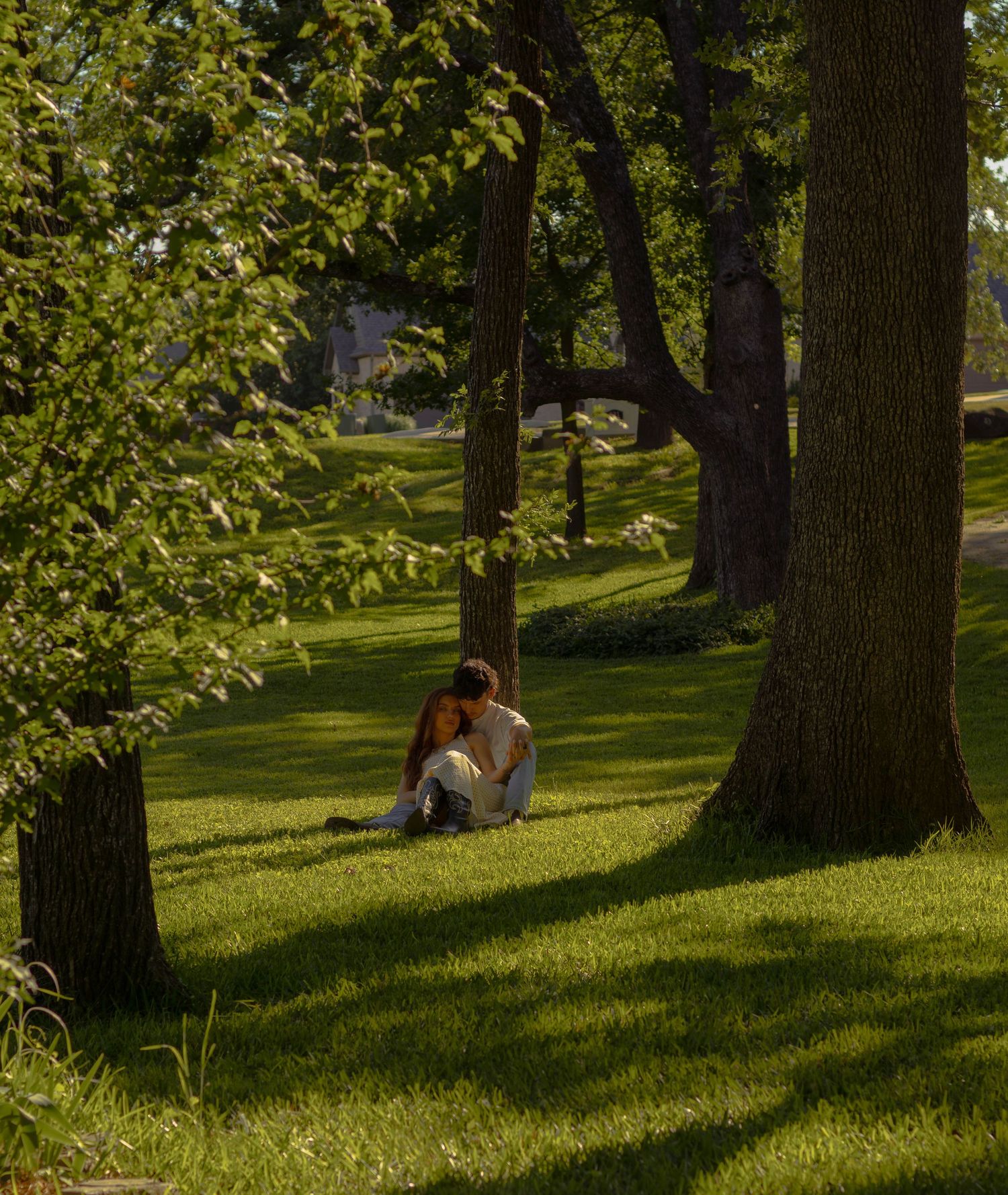 A couple sits together against a tree in a lush green park, enjoying a peaceful moment in dappled sunlight.