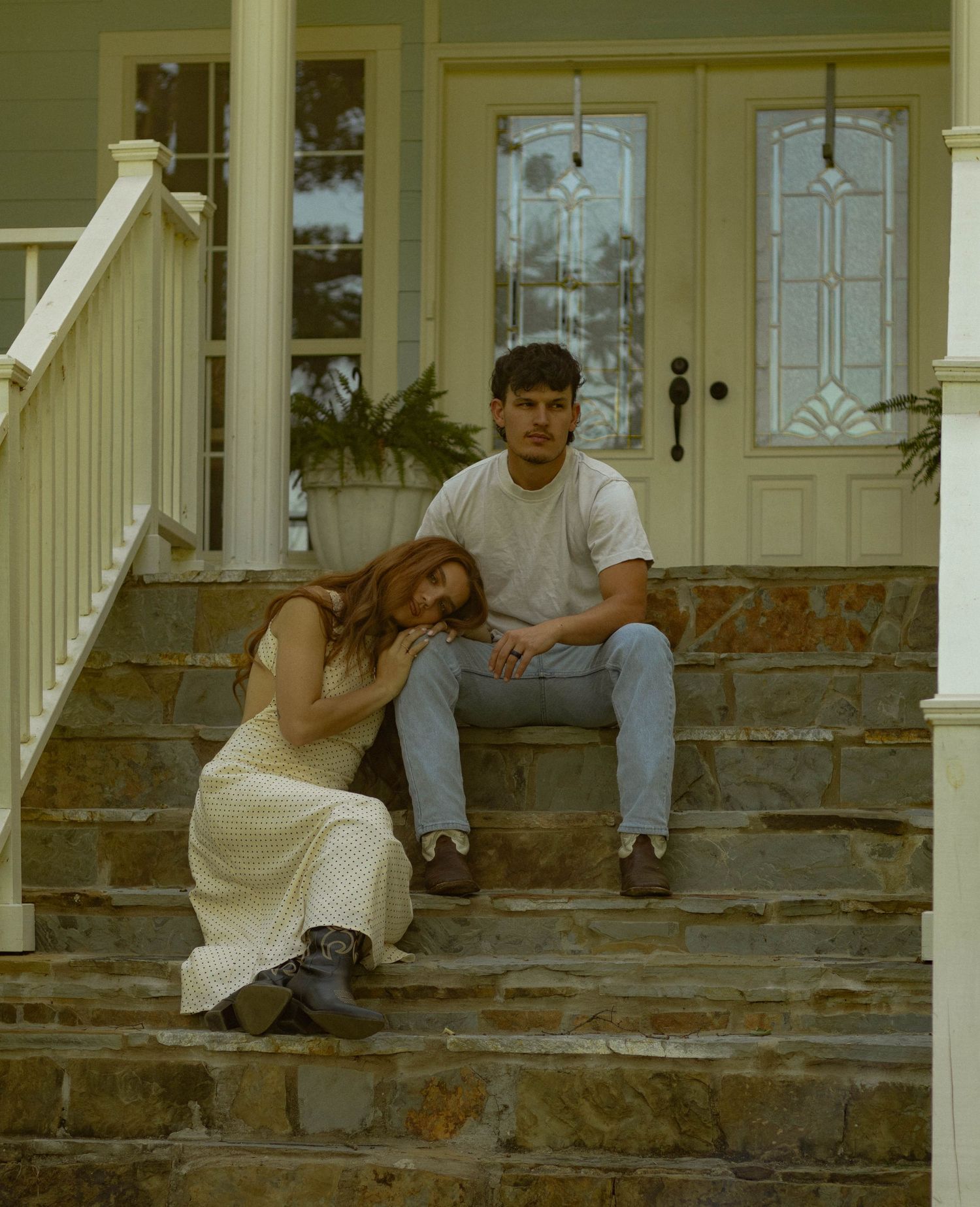A young couple sits on stone steps of a house porch, woman in cream dress leaning on man in white tee and jeans.