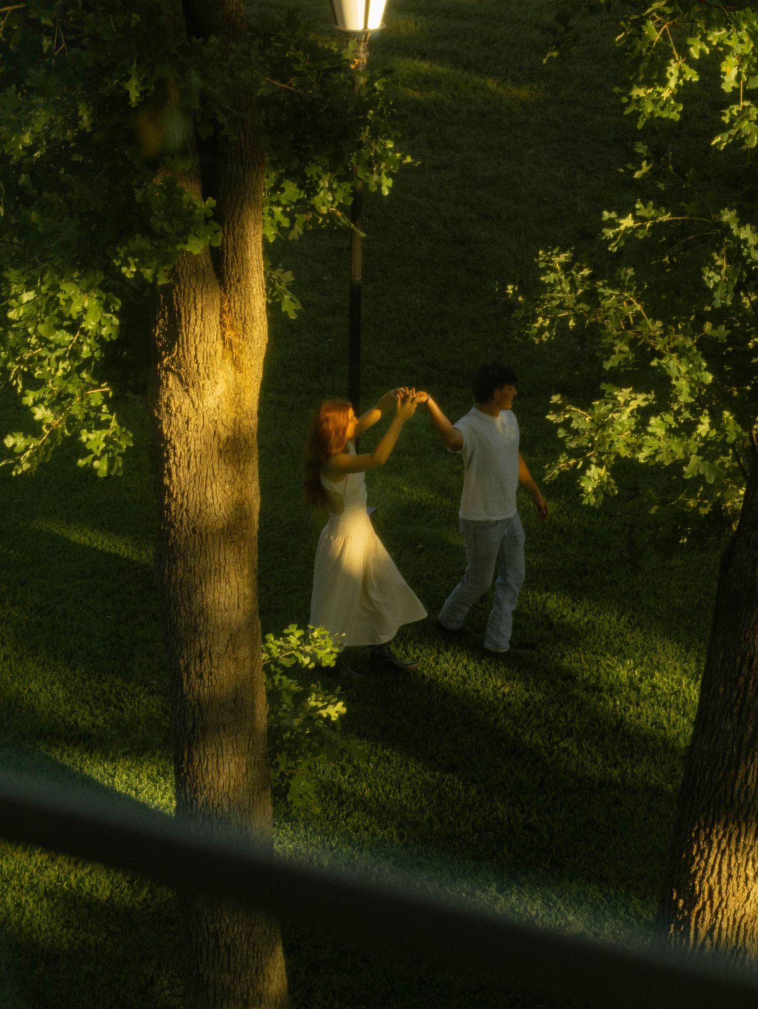 A couple dances romantically under a glowing street lamp surrounded by lush green trees at dusk.