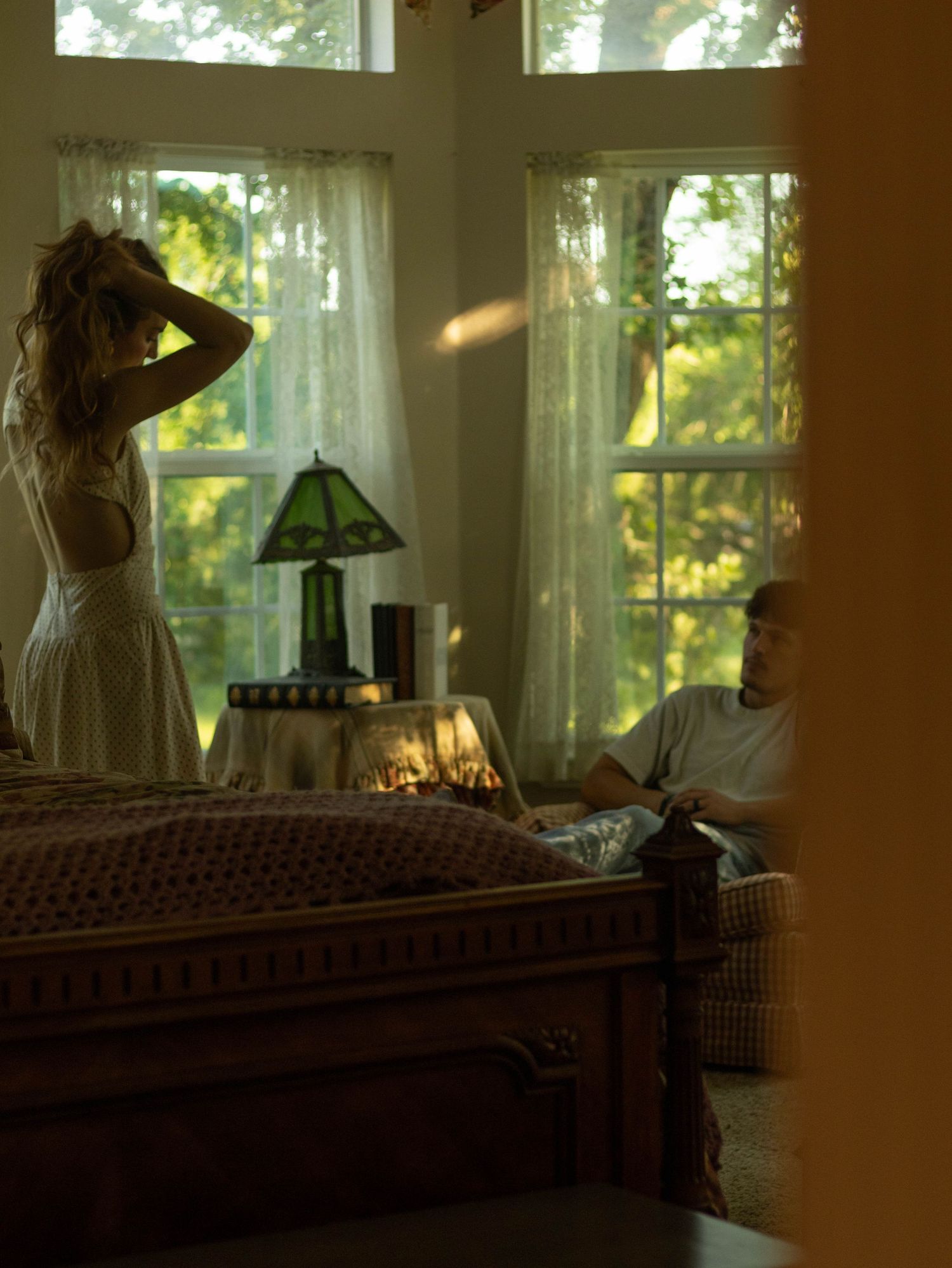 A woman in a white dress stands by sunlit windows while a man reads in a cozy bedroom.