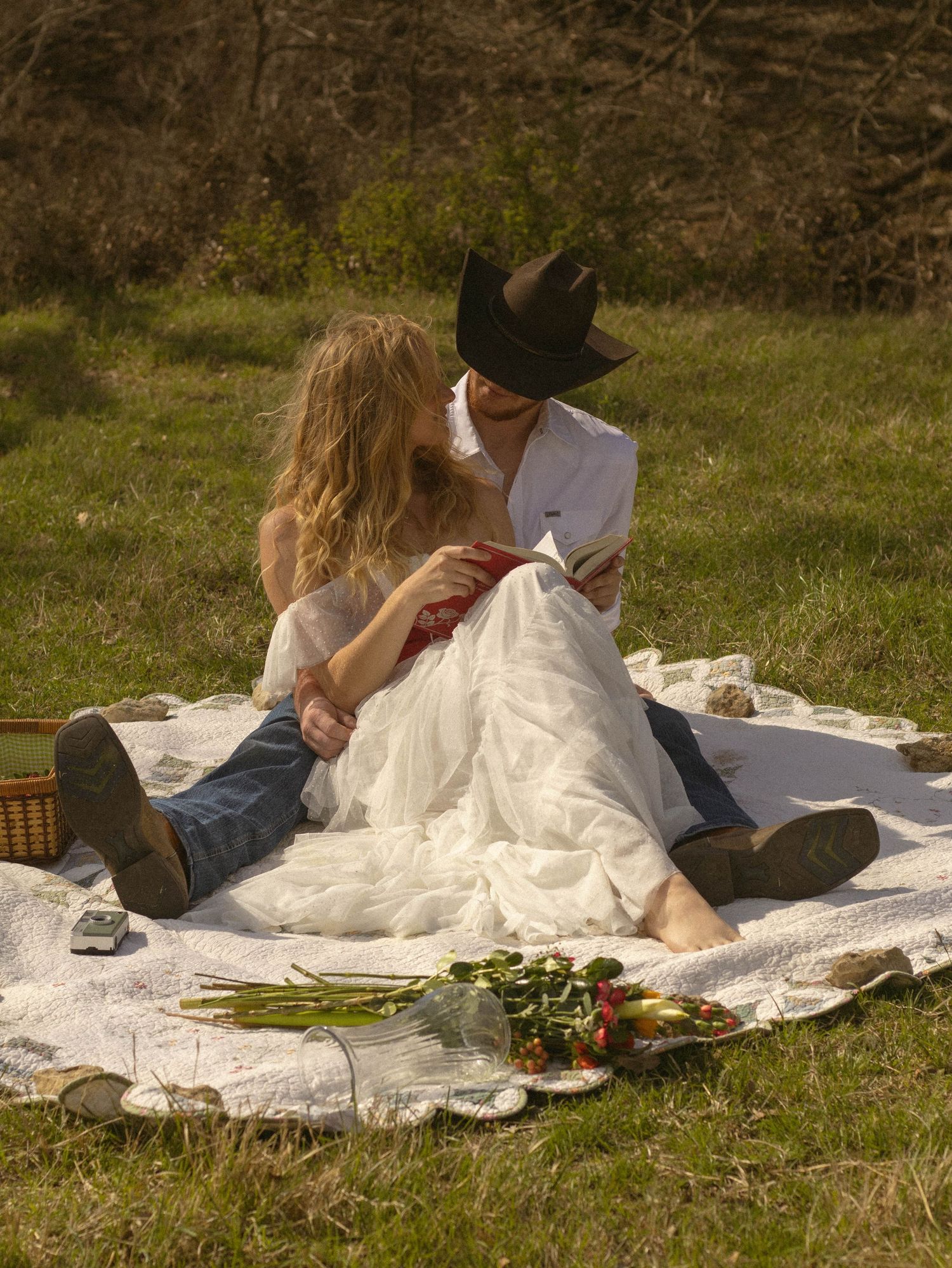A couple relaxes on a white blanket having a romantic picnic in a grassy meadow on a sunny afternoon.