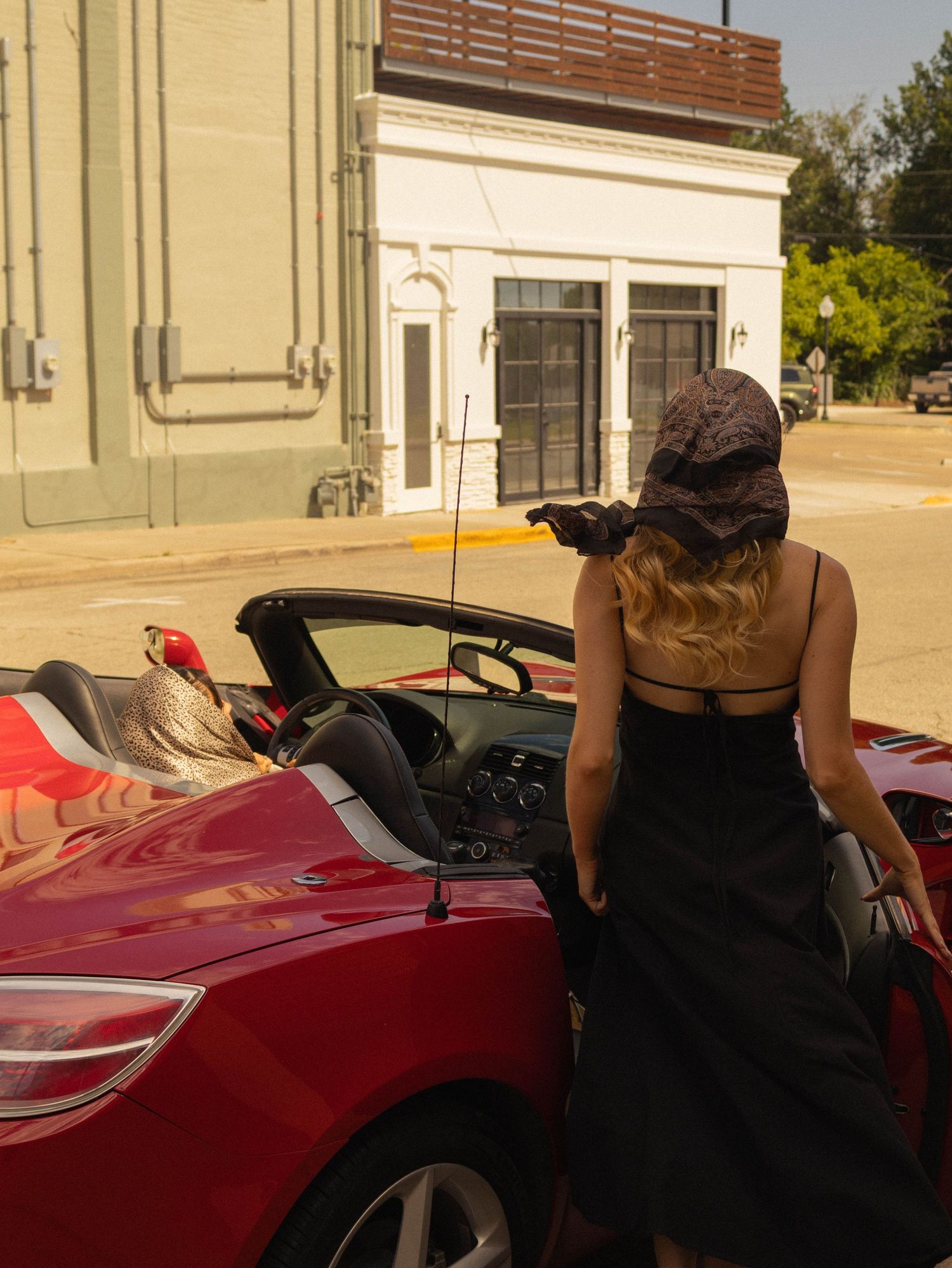A woman in a black dress and hat leans against a red convertible sports car on a sunny small-town street.
