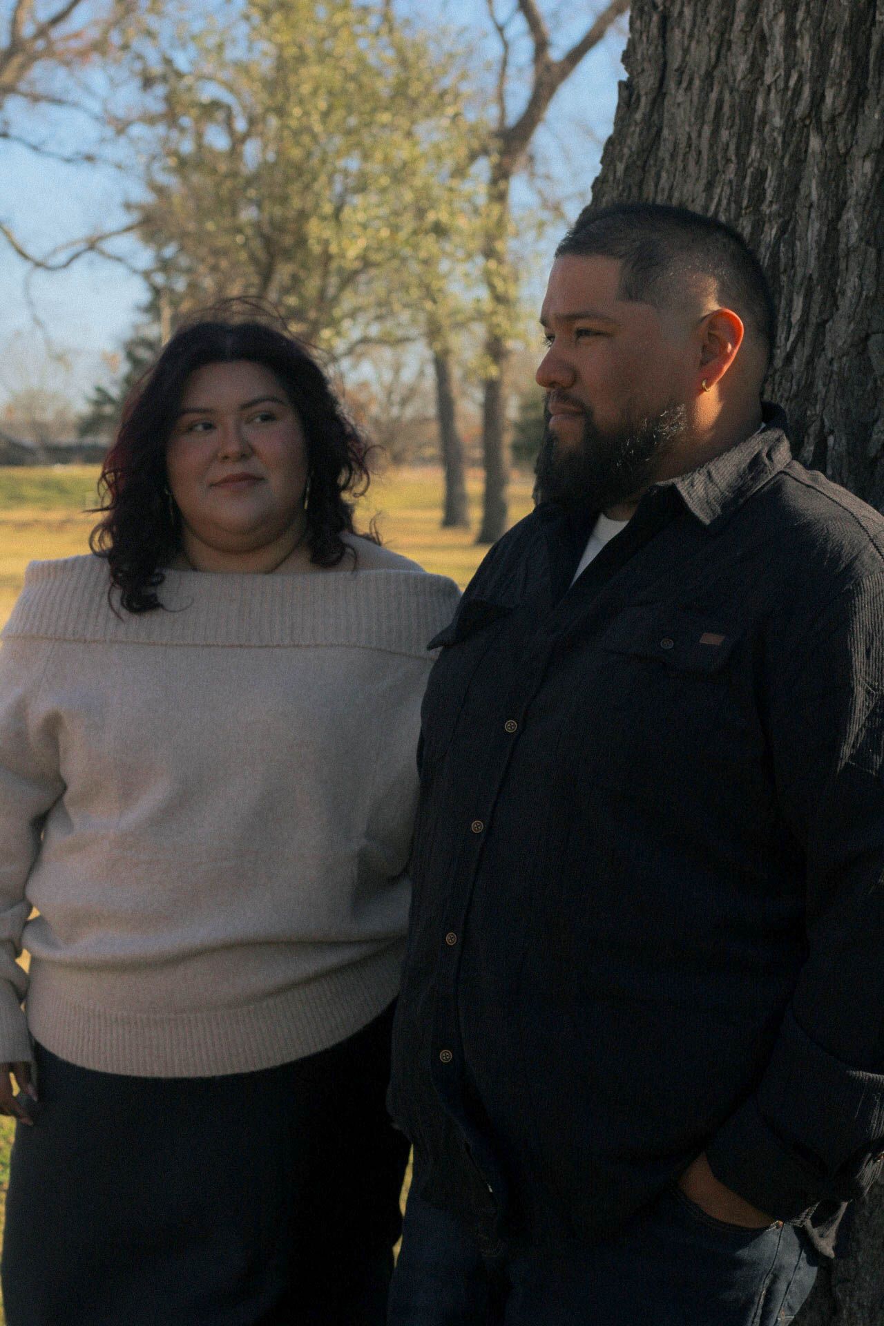 A couple in casual winter attire stands beside a large tree trunk in a park.