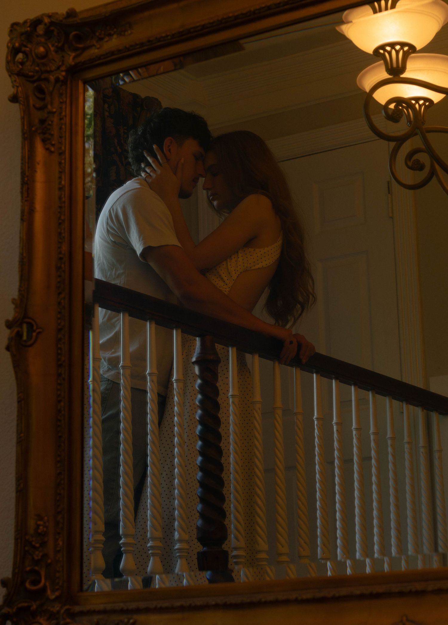 Romantic couple nearly kissing on a balcony reflected in an ornate gold-framed mirror indoors.