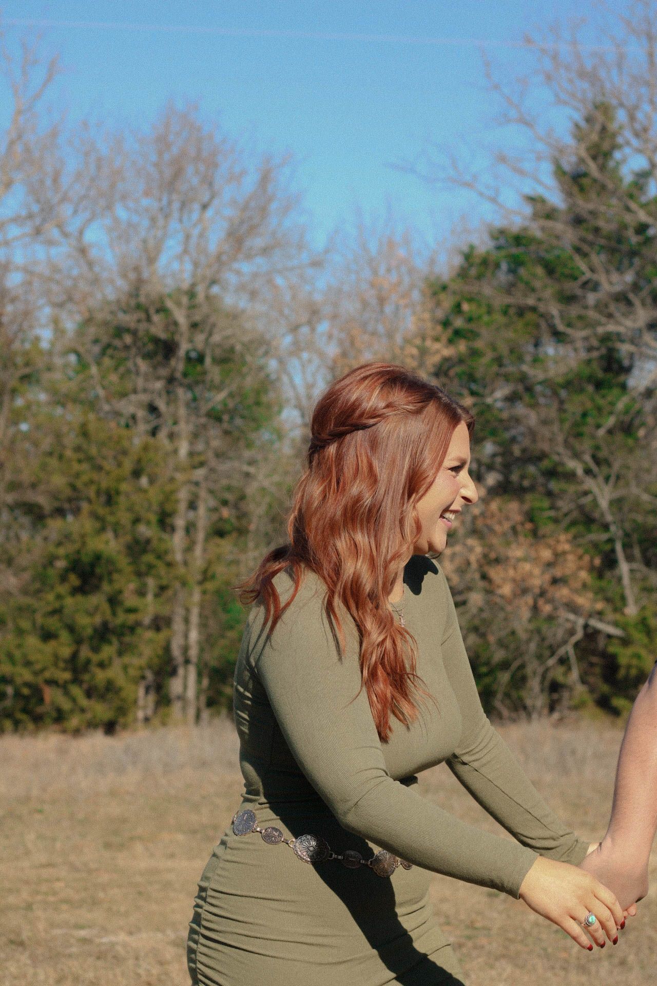 Person in olive green sweater laughs joyfully in an outdoor setting with bare trees.