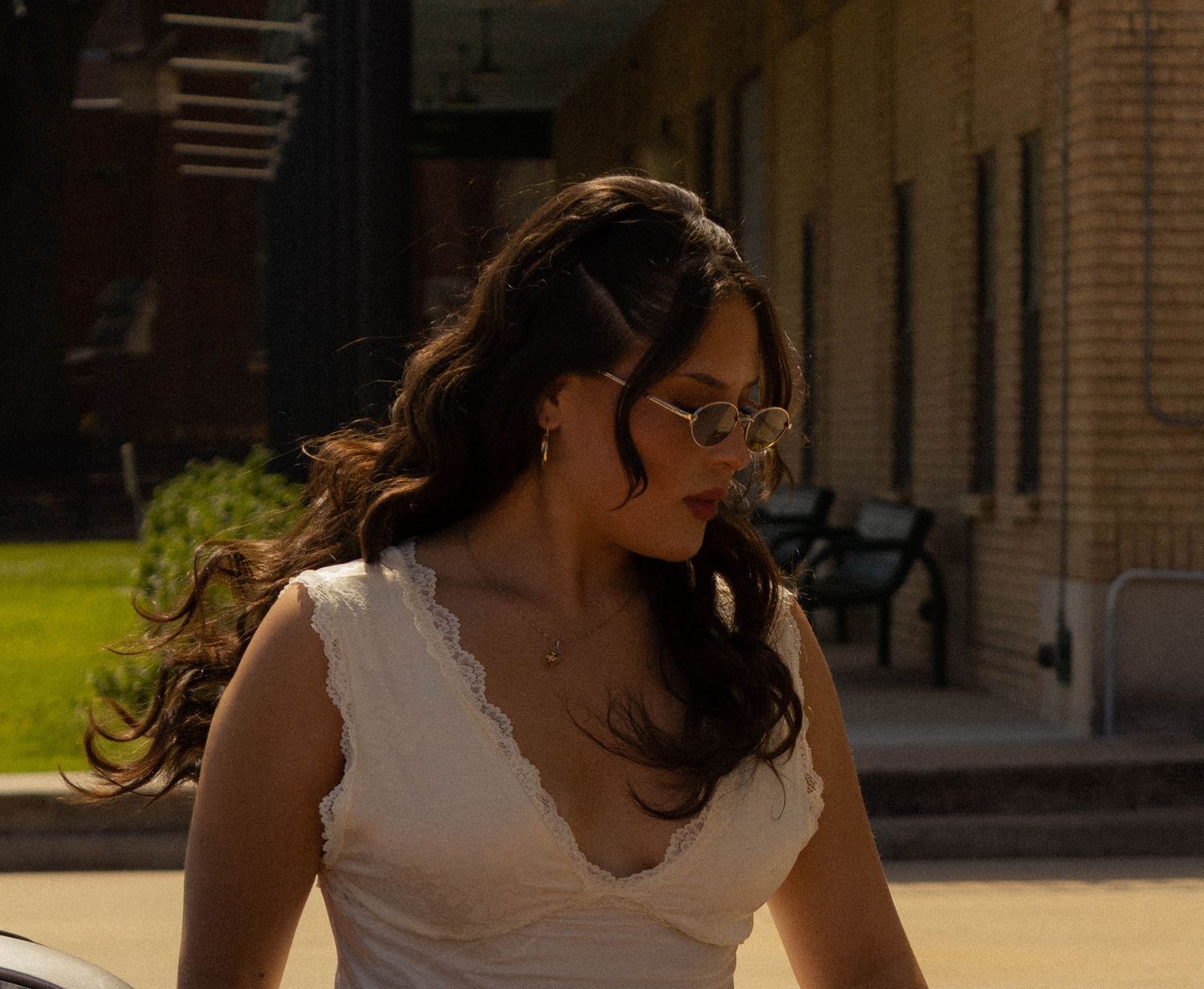 Woman in white lace dress and sunglasses with long dark hair outdoors near brick building.