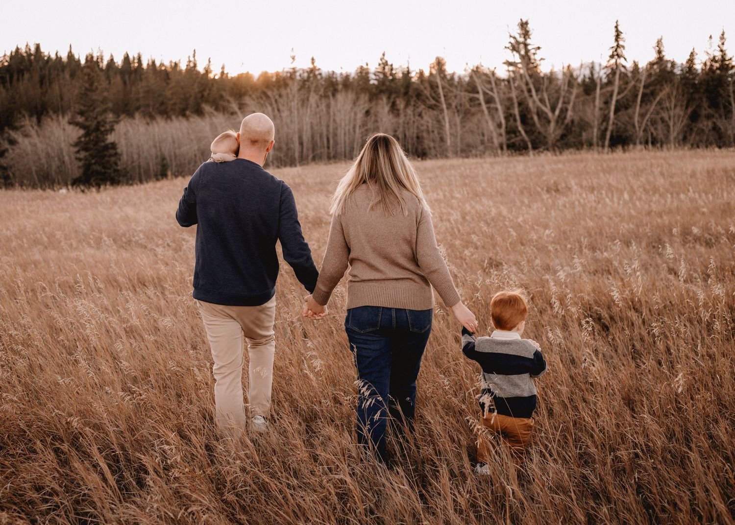 A family walks hand in hand through a golden Calgary, AB meadow at sunset.