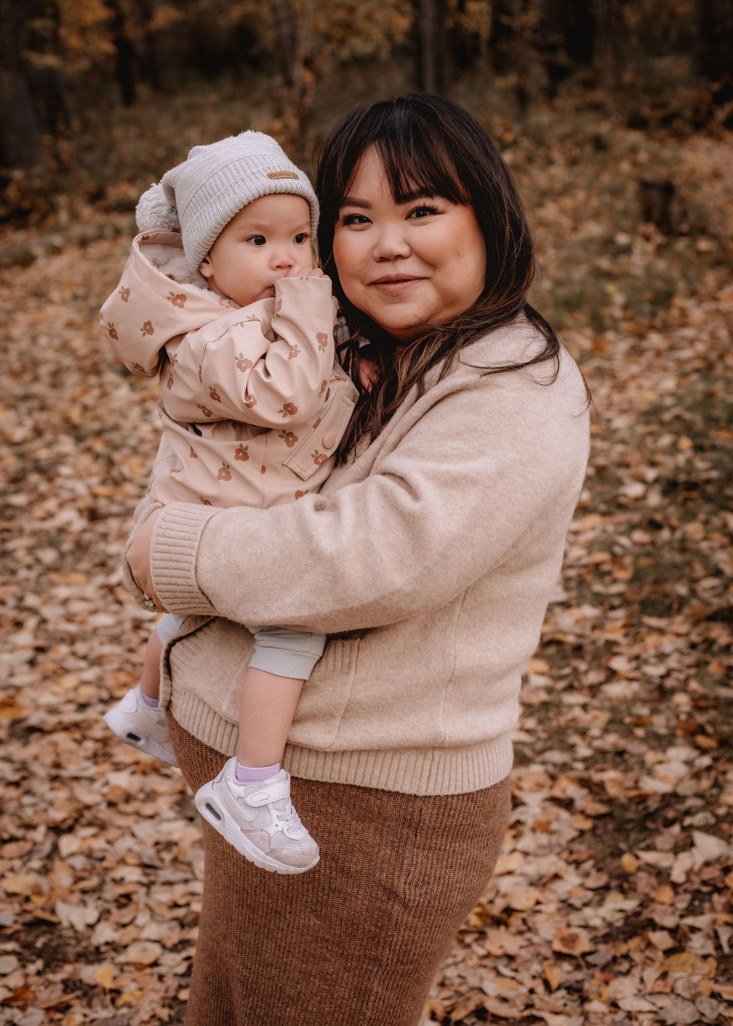 A smiling woman holds her baby in a beige outfit among autumn leaves in a forest setting for lifestyle photoshoot