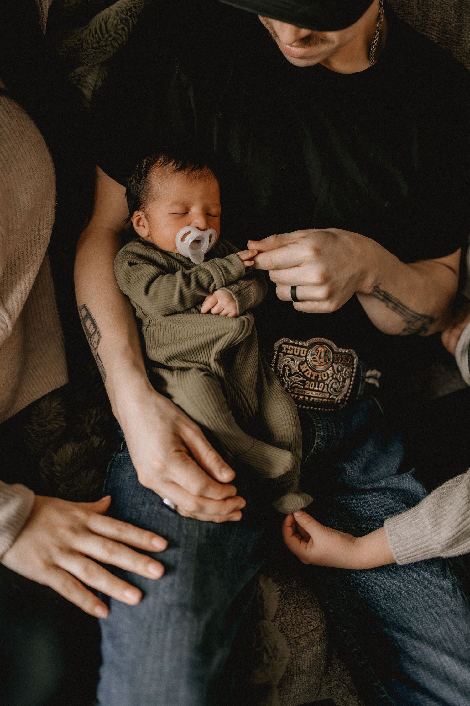 Tattooed father holding sleeping newborn baby in olive green onesie with pacifier on his lap.
