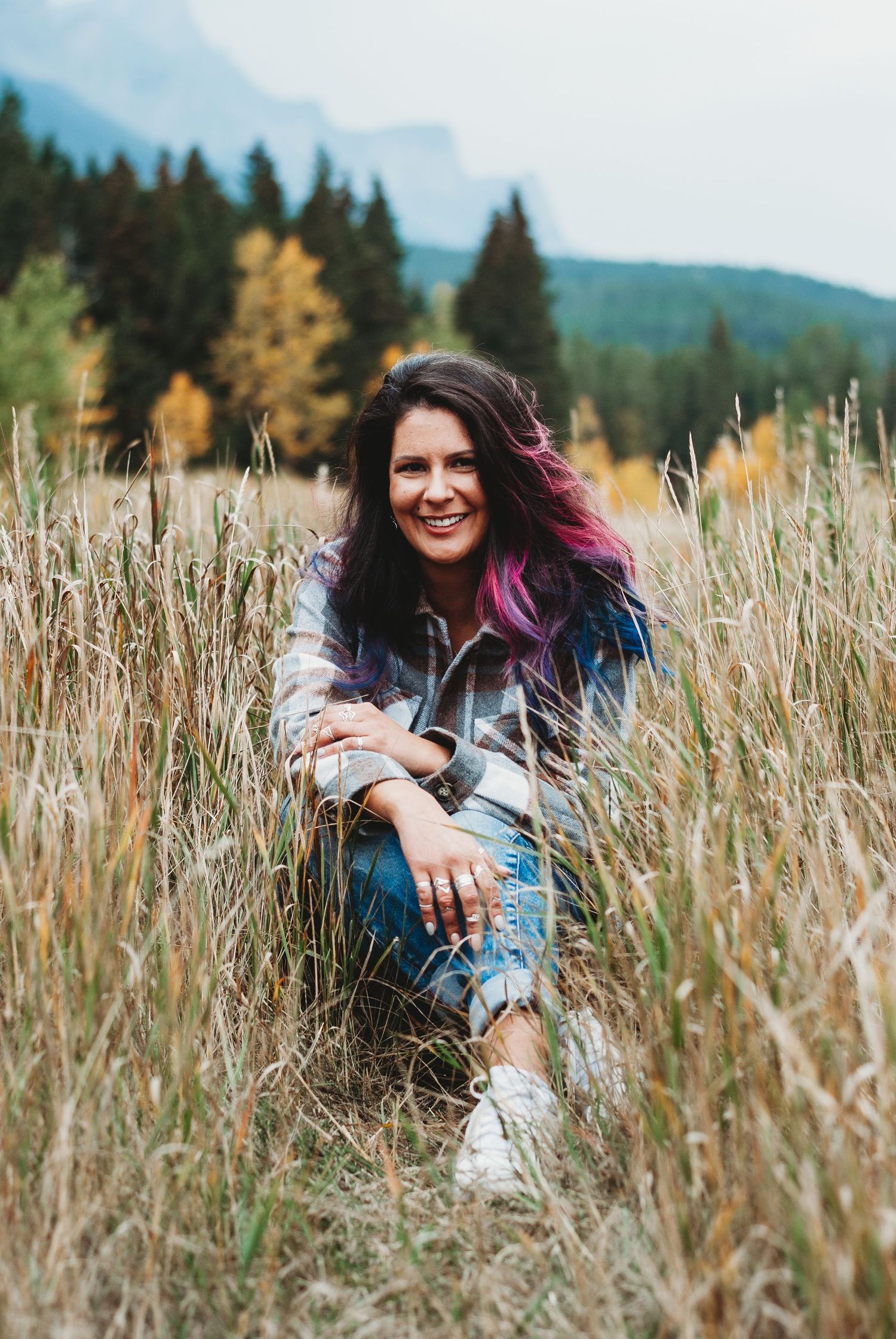 Woman with purple-tipped hair smiling in a golden field, wearing plaid shirt and jeans, with Rocky Mountains in background.