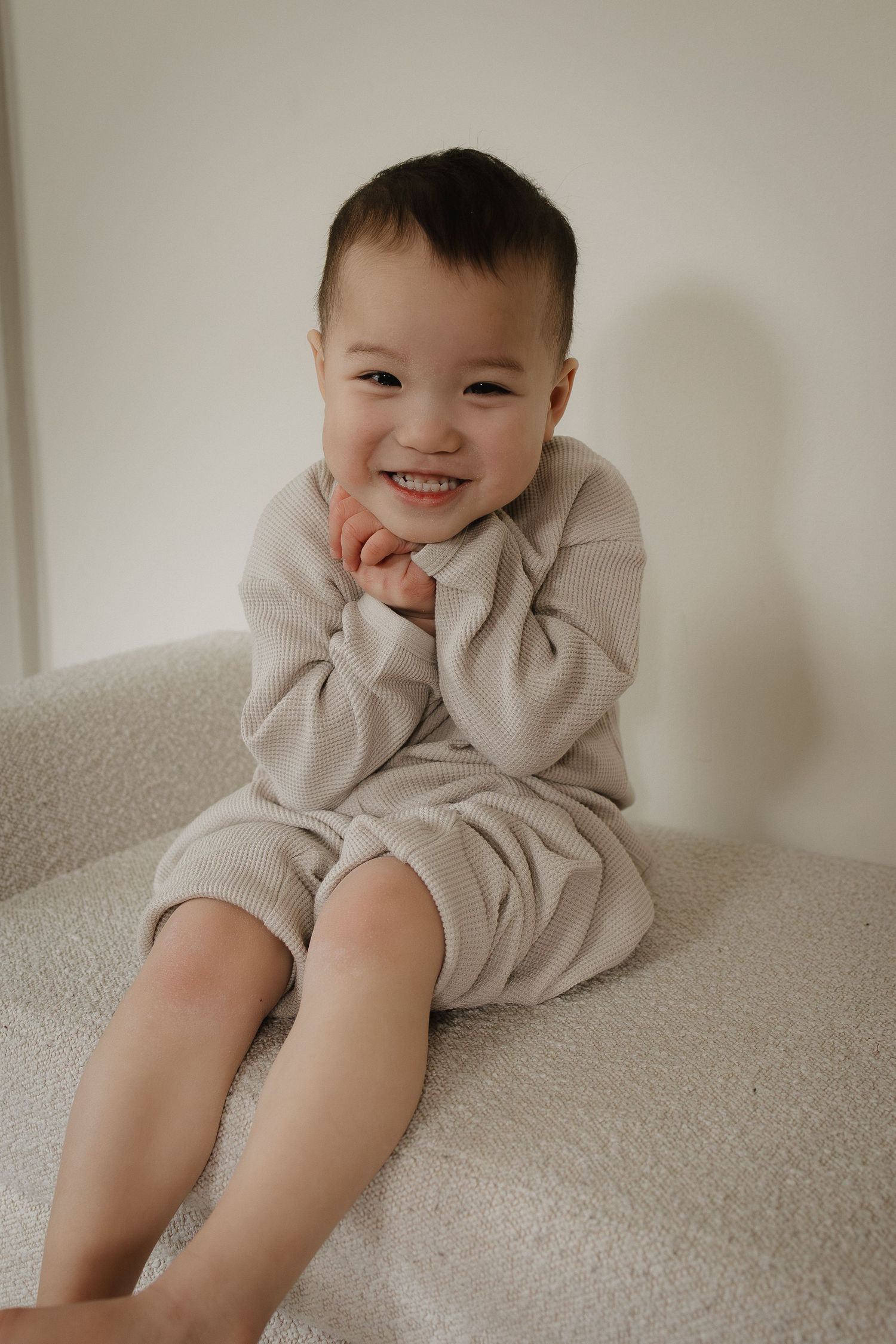 Calgary family photographer captures a smiling toddler in beige outfit sitting on white chair with hands under chin.