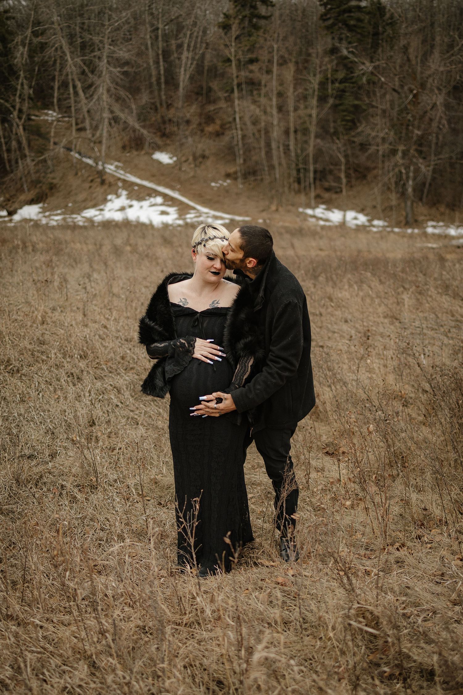 Couple in black outfits poses in a field during a winter maternity photoshoot with snowy trees in background.