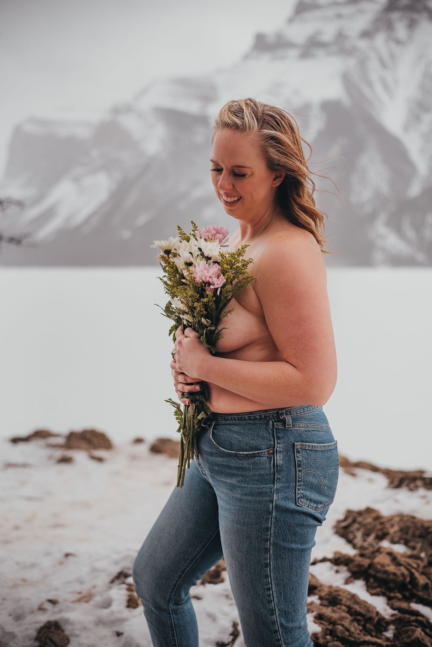 Woman holding pink flower bouquet outdoors in snowy Banff mountain landscape wearing blue jeans, smiling.