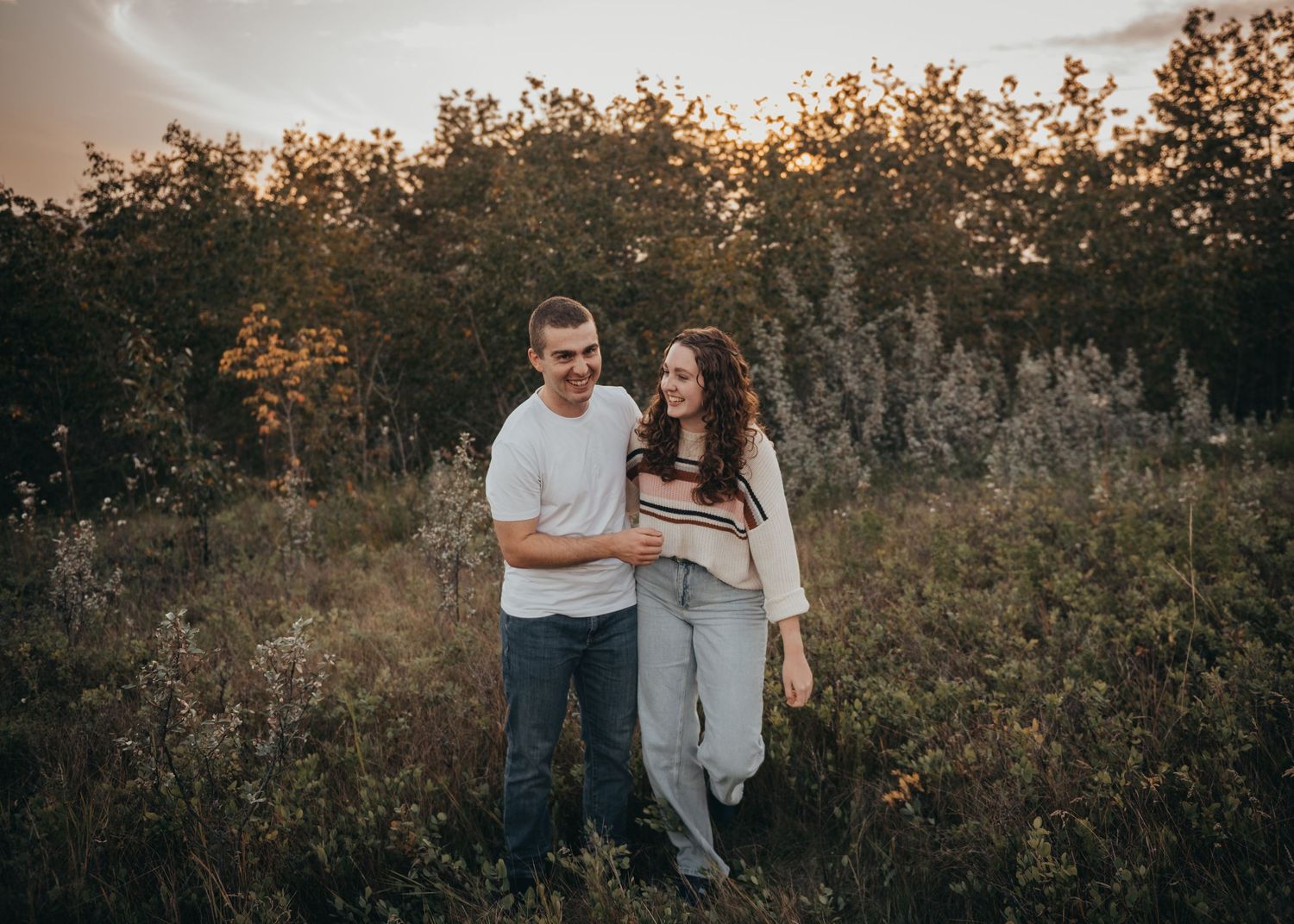 Siblings smiling and walking together through a golden Calgary field at sunset, surrounded by wildflowers and trees.