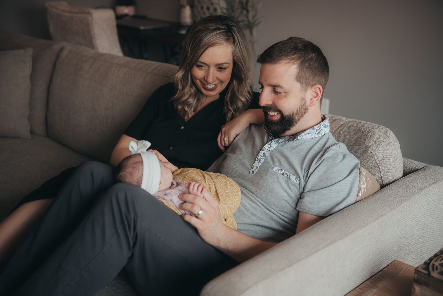 A happy Calgary couple sitting on a cozy sofa, lovingly gazing at their newborn baby in a warm home setting.
