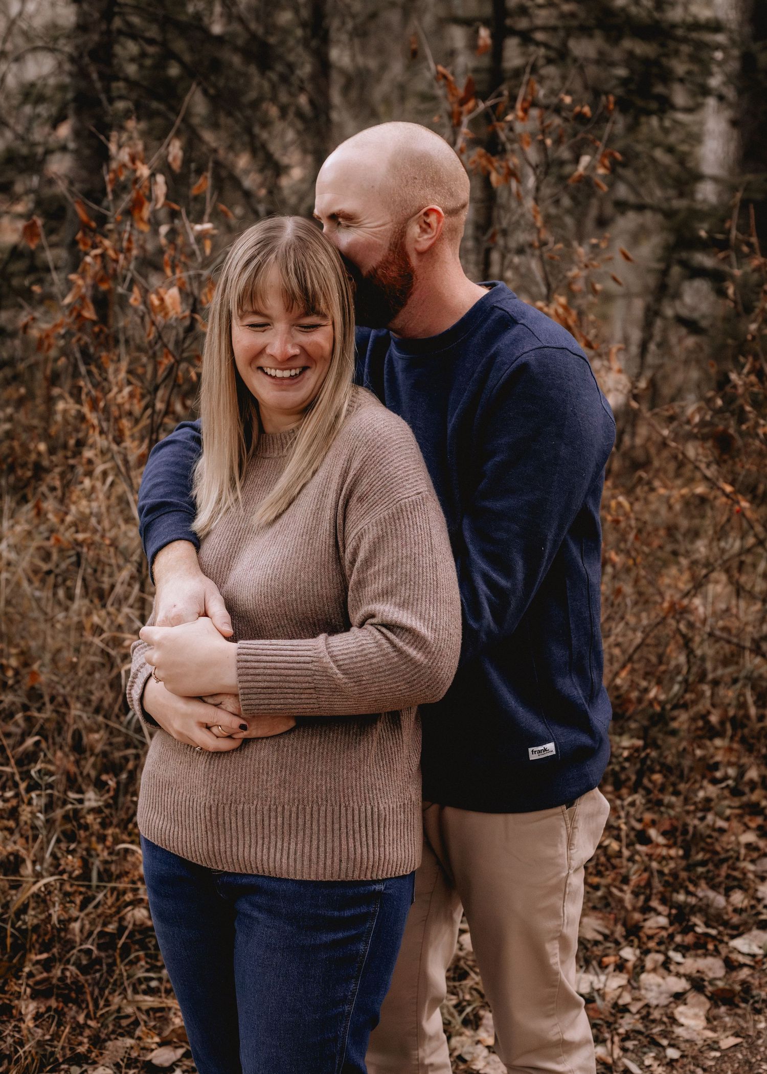 A smiling couple embraces outdoors in autumn Fish Creek Park, AB woods, man in navy sweater hugging woman in tan sweater from behind.