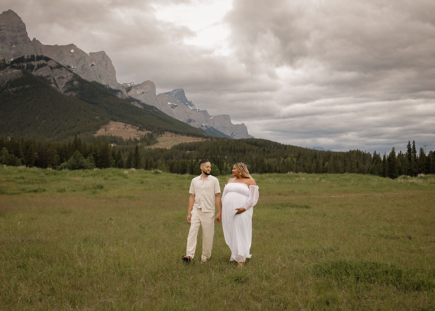Pregnant mom and dad holding hands in front of mountain in open field with soft natural light spilling over them in Calgary