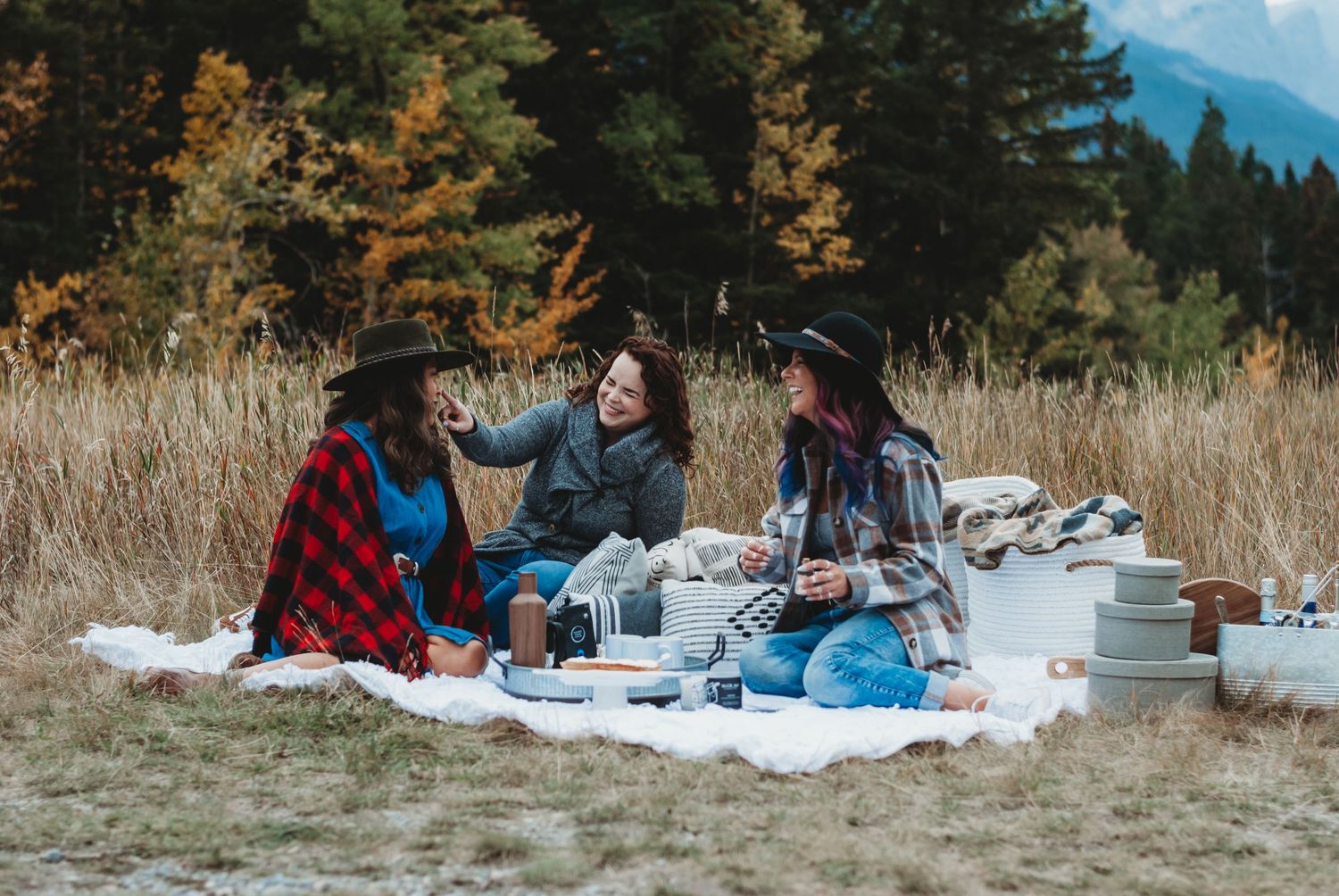 Three women enjoy an outdoor picnic on a white blanket in a scenic mountain meadow surrounded by autumn trees.