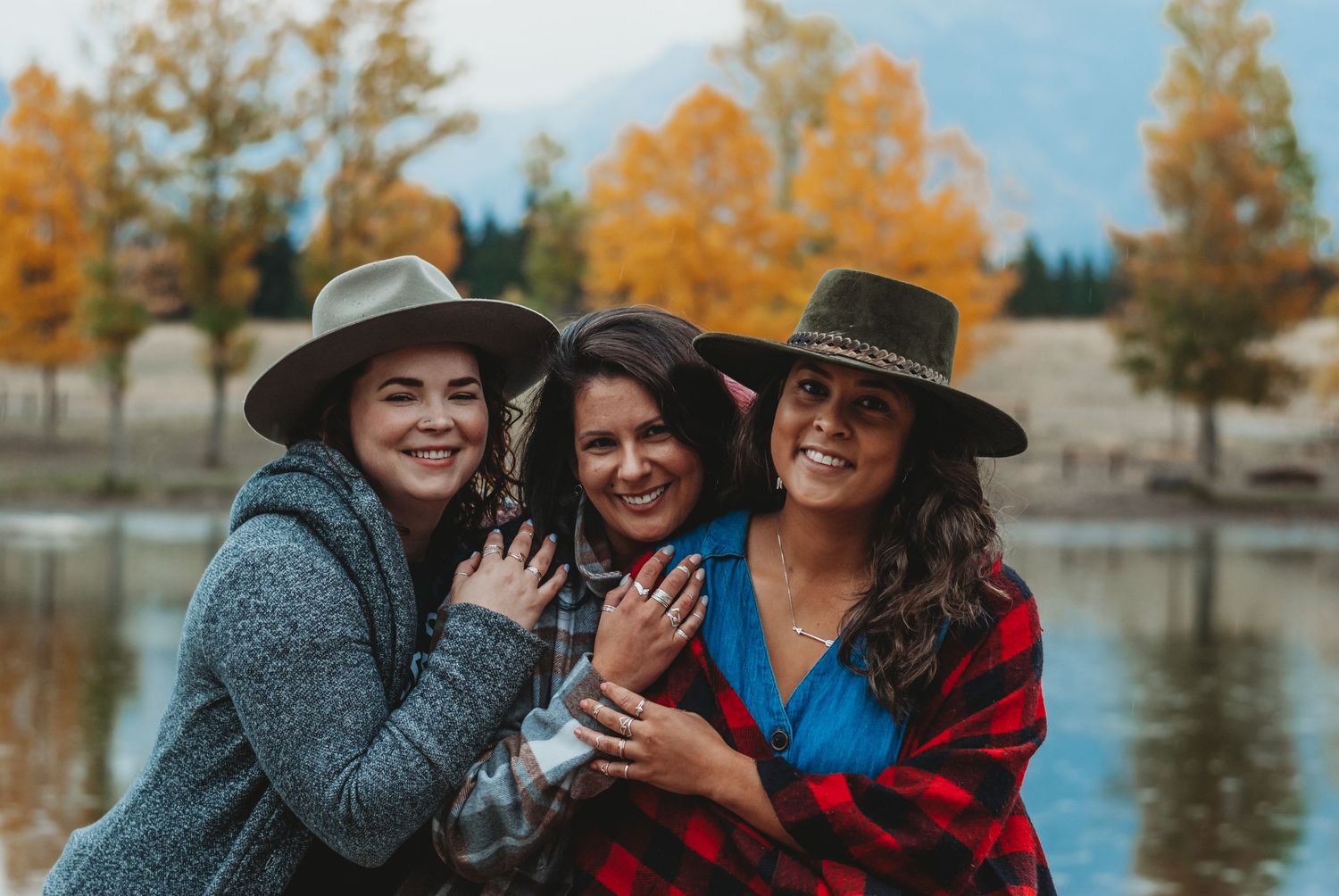 Three women in wide-brim hats smile together near Quarry Lake with autumn trees and mountains in the background.