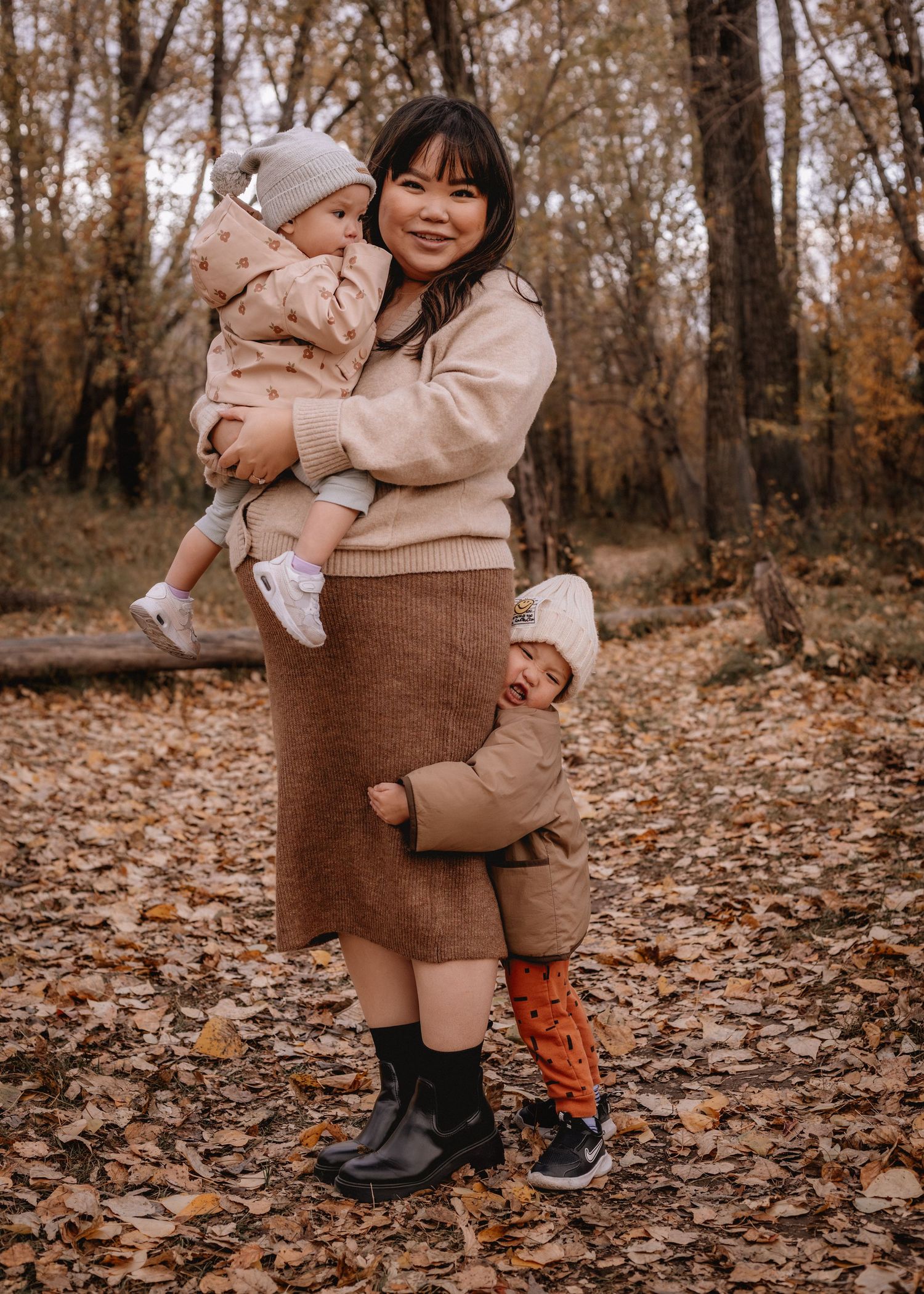 A woman holds a baby while a toddler clings to her skirt among autumn leaves in a wooded Calgary setting.