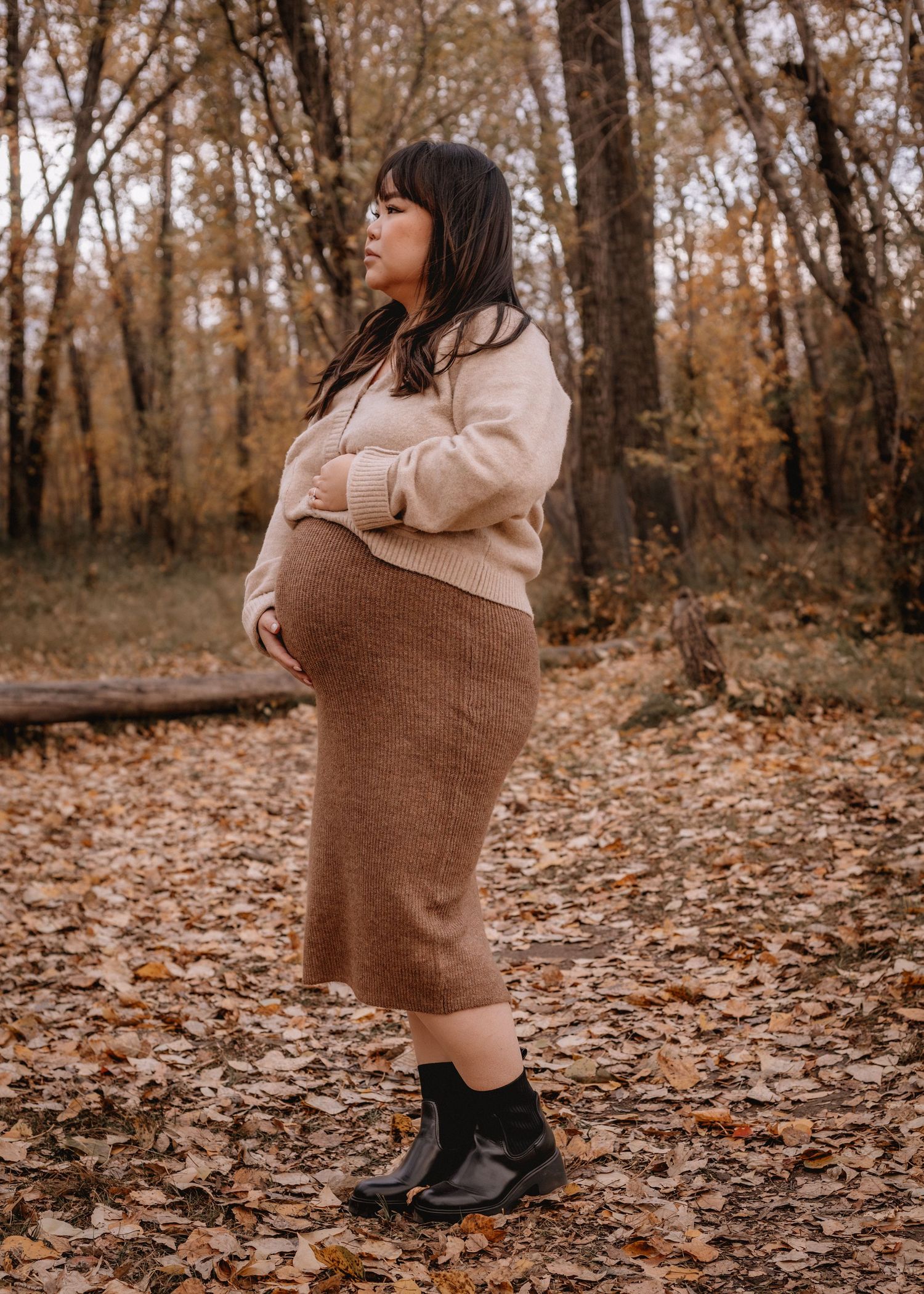Pregnant woman in beige sweater and brown midi skirt poses in autumn forest with fallen leaves in Calgary, AB.