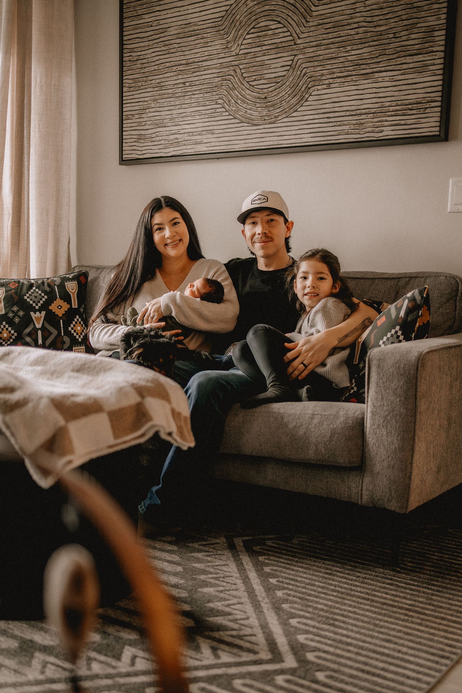 Family of four sitting on a gray couch, parents holding a newborn and young daughter, in a cozy modern living room.