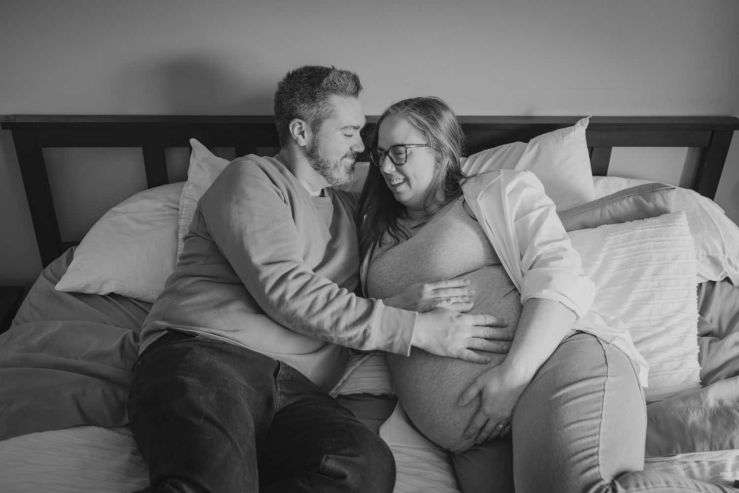 A candid black and white maternity session photo of a couple sharing a tender moment on a bed, with the woman visibly pregnant.