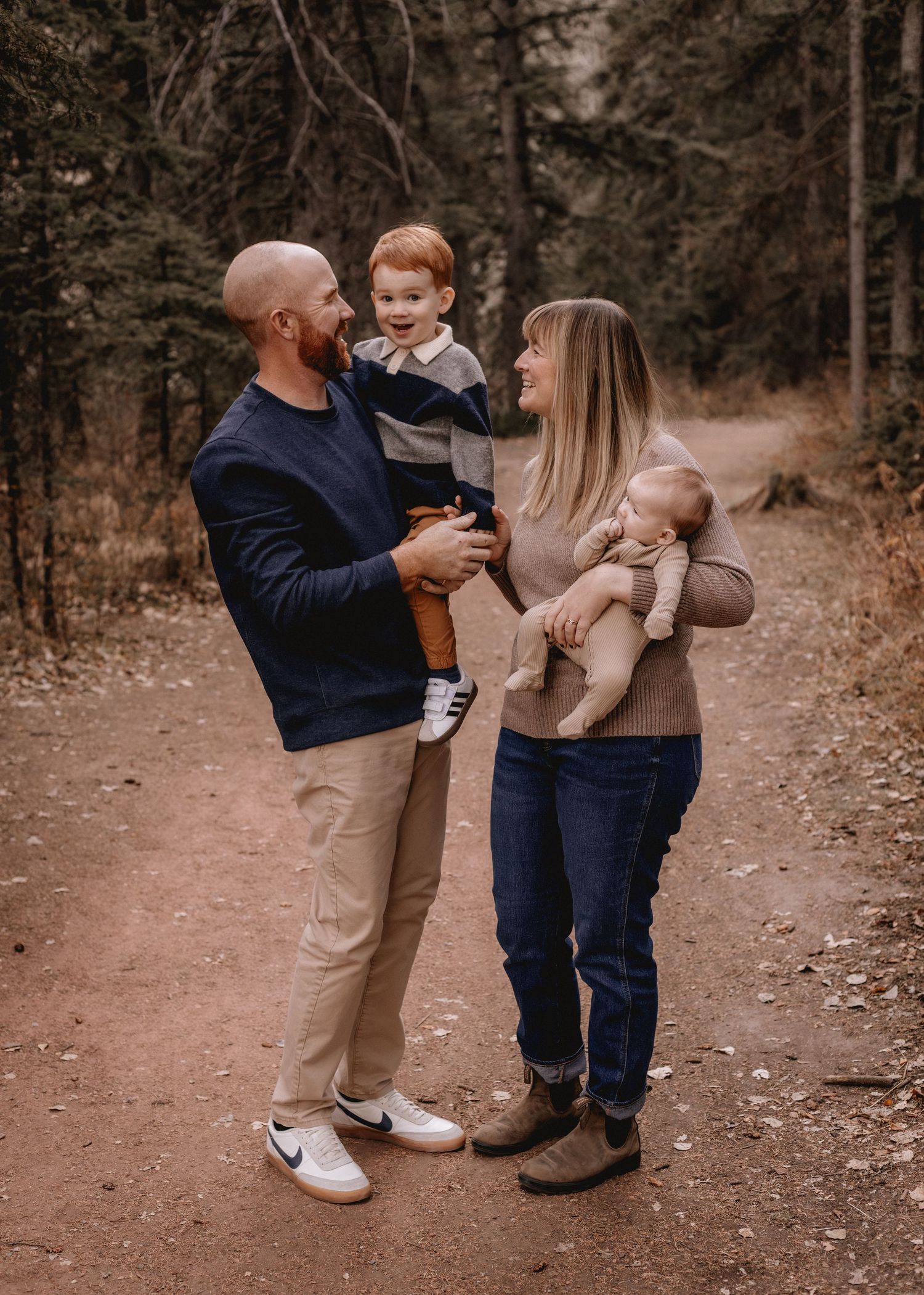 Family of four posing on a forest trail, parents holding two young children, surrounded by autumn trees.