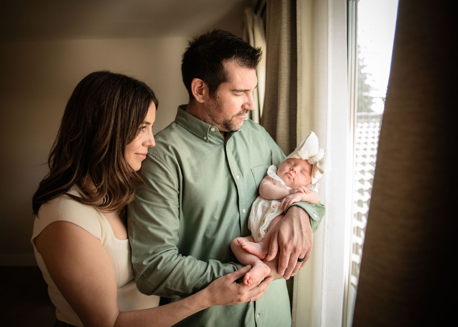 Parents gazing lovingly at their newborn by a sunlit Calgary, AB window.