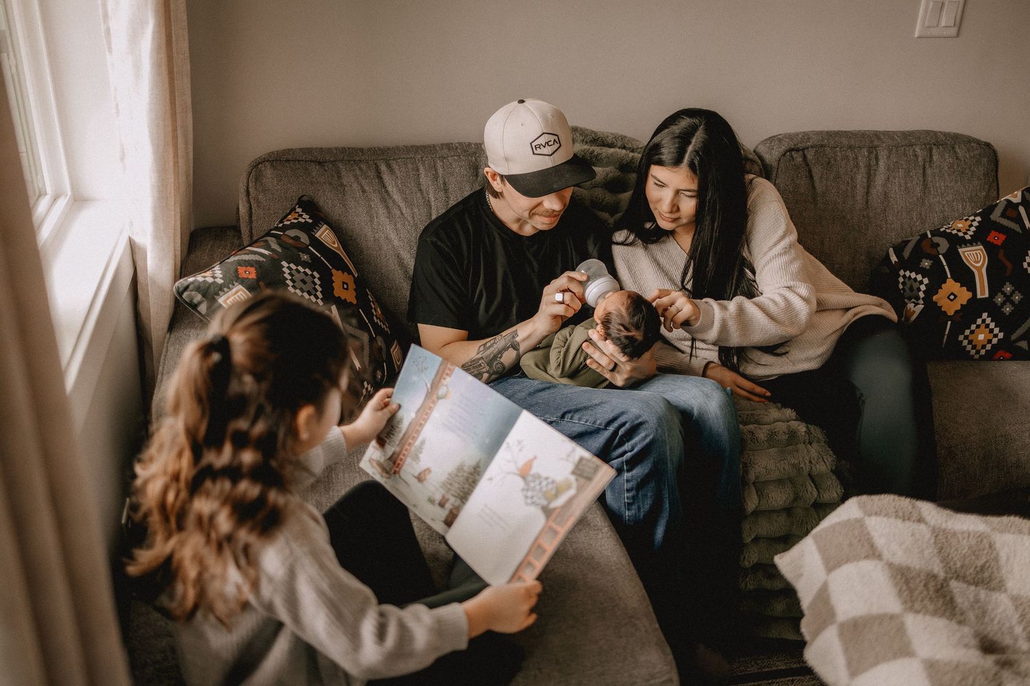 Family sits on couch reading a picture book together with newborn baby in a cozy living room in Calgary, AB.