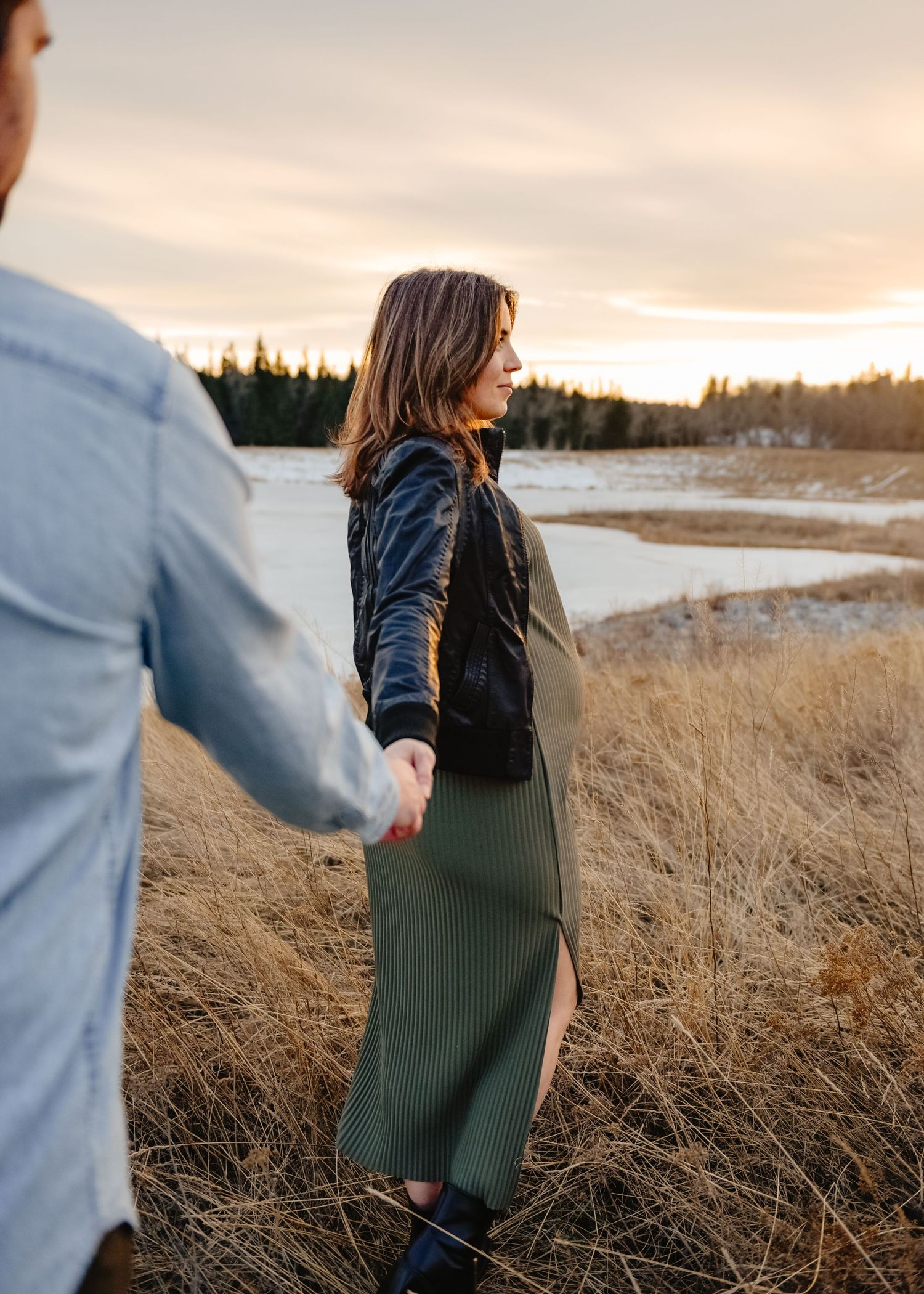 Couple holding hands in a golden field at sunset in Calgary, woman wearing olive midi dress and velvet jacket.