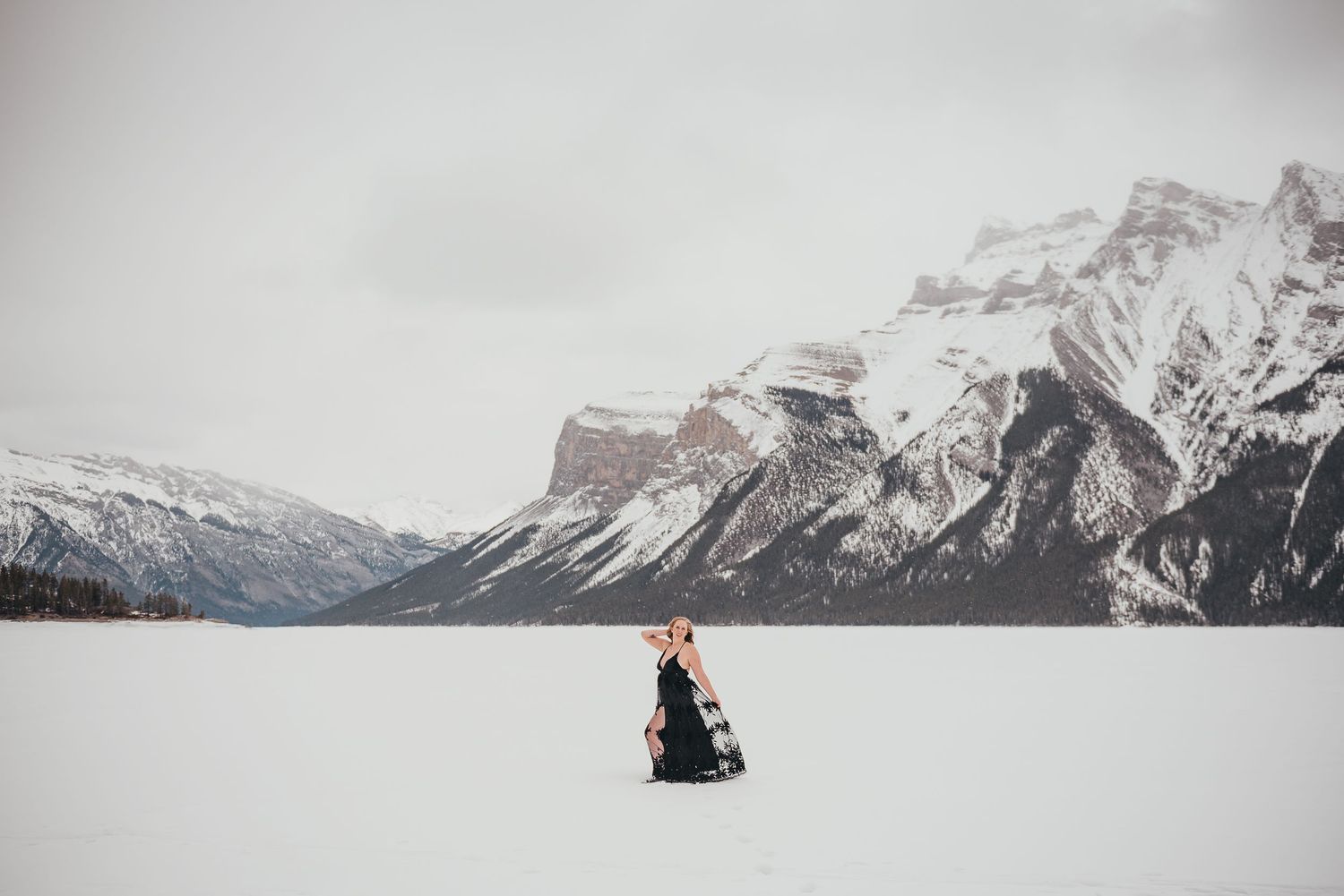 Woman in black dress stands on frozen lake with snow-capped mountains in background during winter.