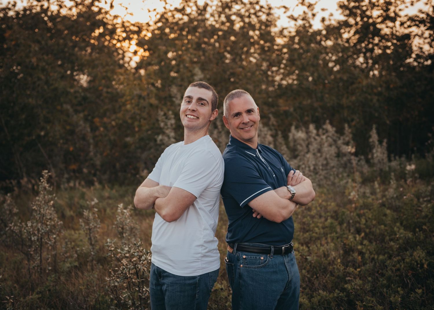 Two men standing back-to-back with arms crossed smiling in a golden hour outdoor field setting.