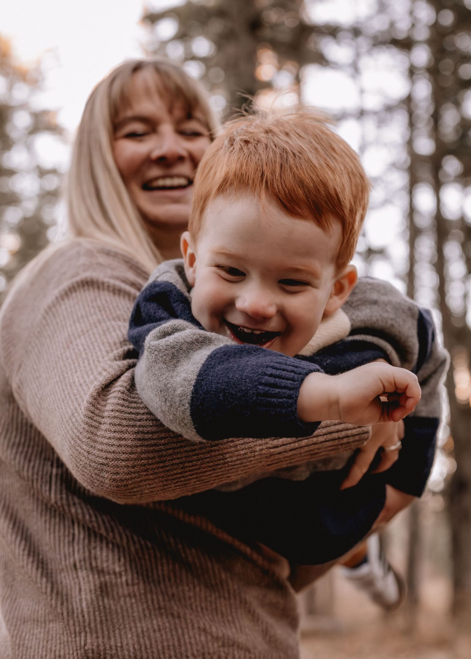 A smiling woman holds a laughing red-haired toddler boy outdoors in a forest setting for family portraits.