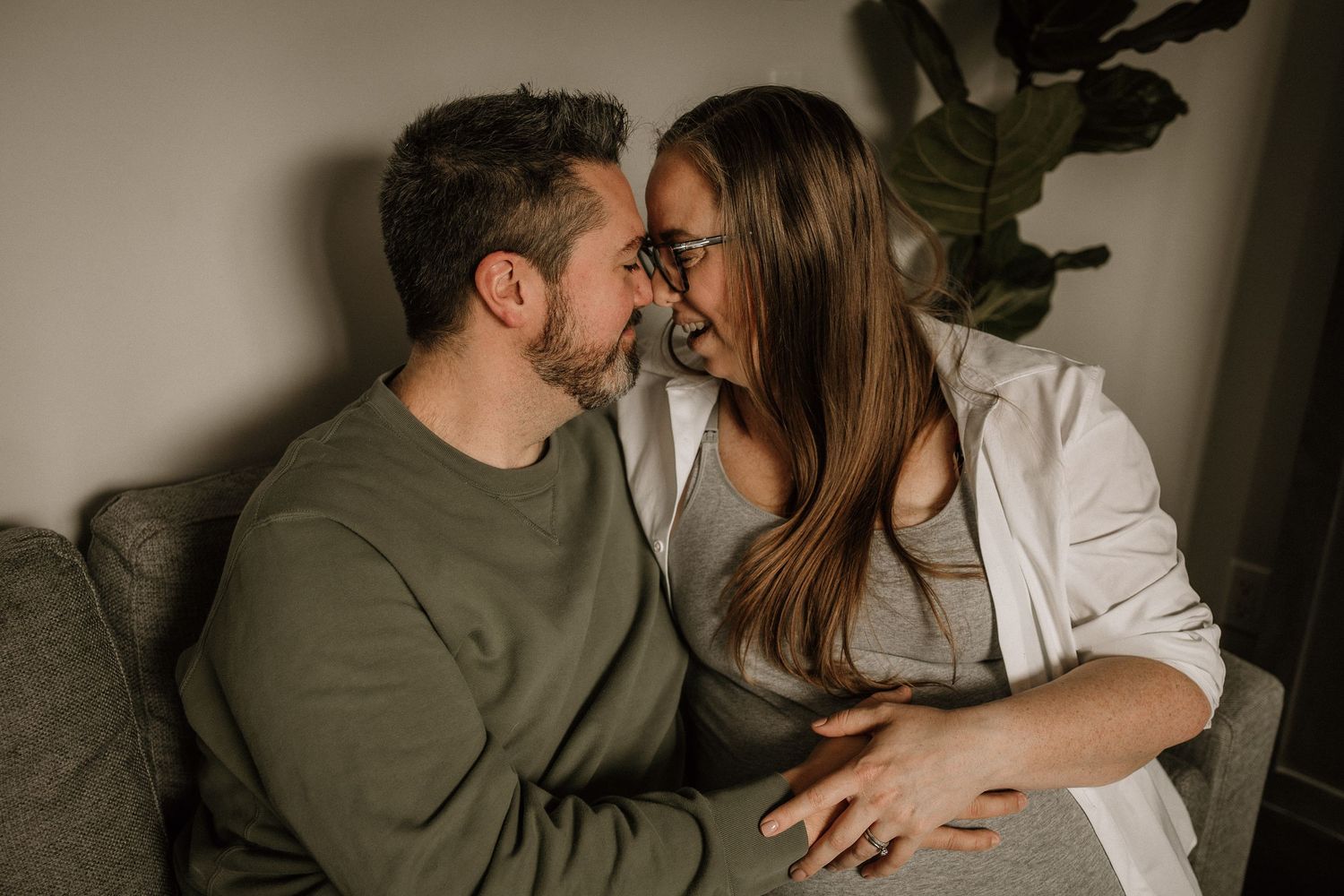 Couple sharing a tender moment on a couch, woman wearing glasses and white shirt, man in green sweater - maternity photography in Calgary.