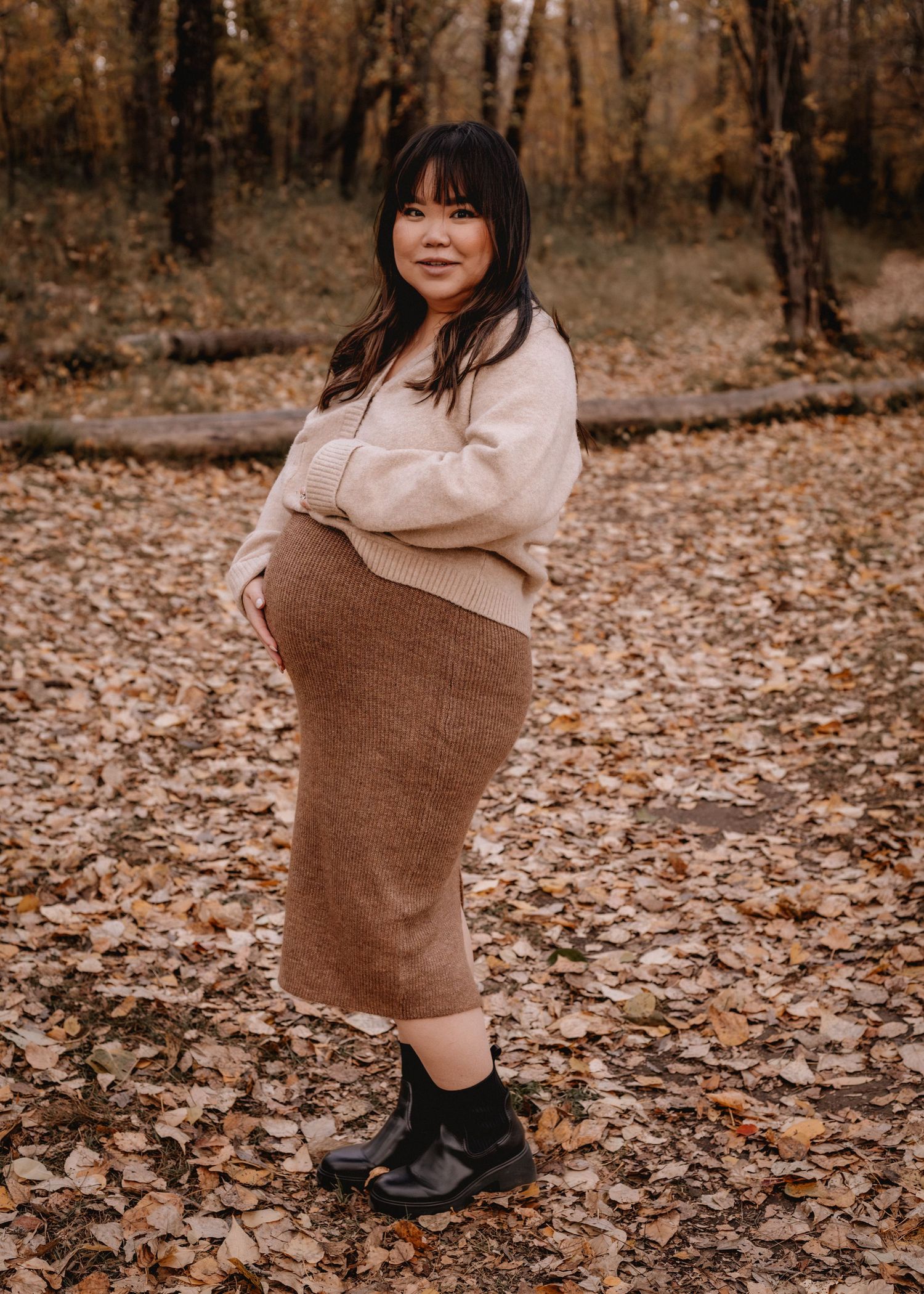 Pregnant woman in beige sweater and brown knit skirt poses in outdoor Calgary, AB among autumn leaves in a forest setting.