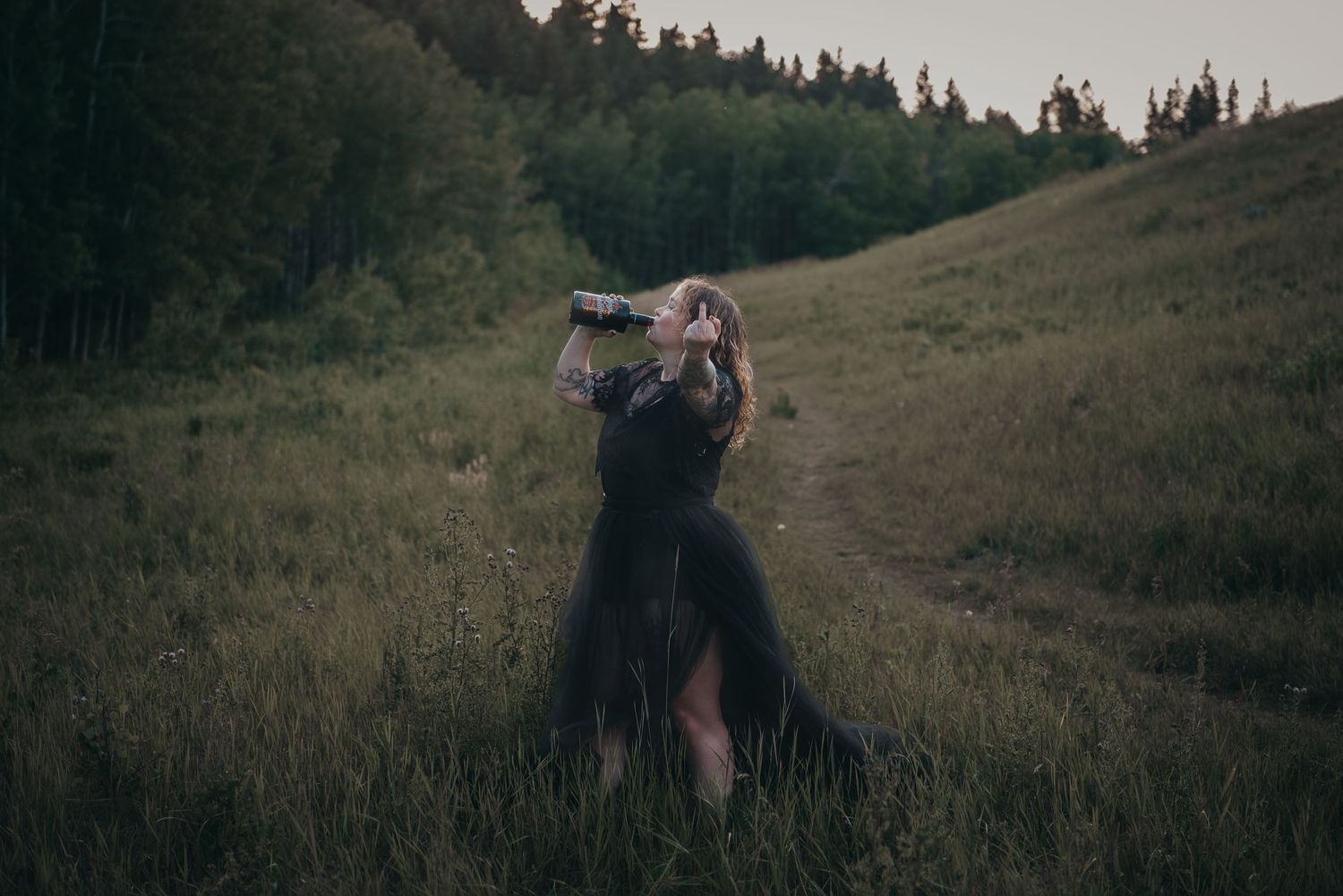 Calgary woman in black dress poses dramatically in a grassy meadow at dusk with forest hills in the background.