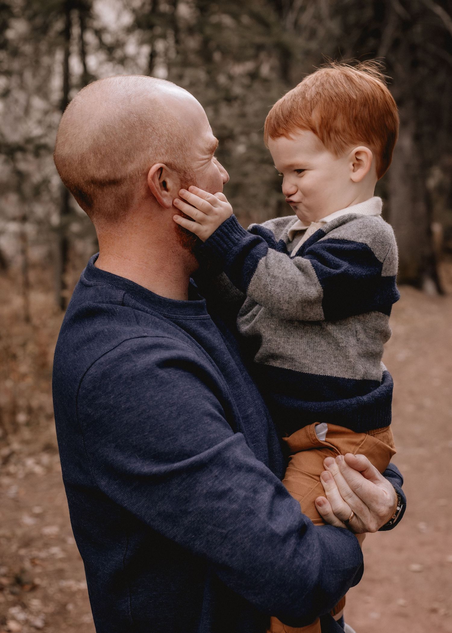 A bald man holds a red-haired toddler boy in a Calgary forest, as the child gently touches his face.