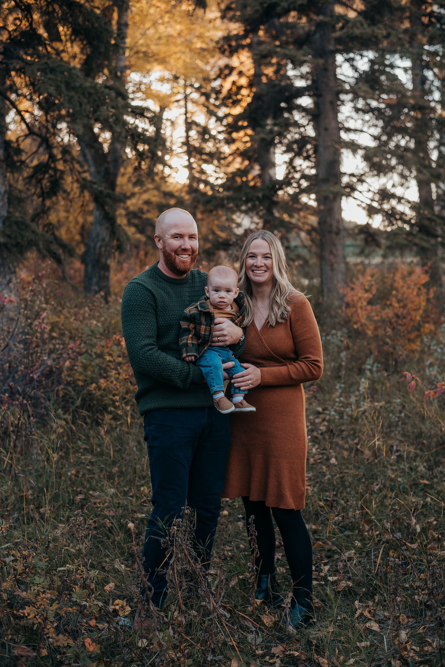 A smiling family of three poses in Bowness Park, AB, parents holding their baby, wearing cozy fall outfits in warm tones.