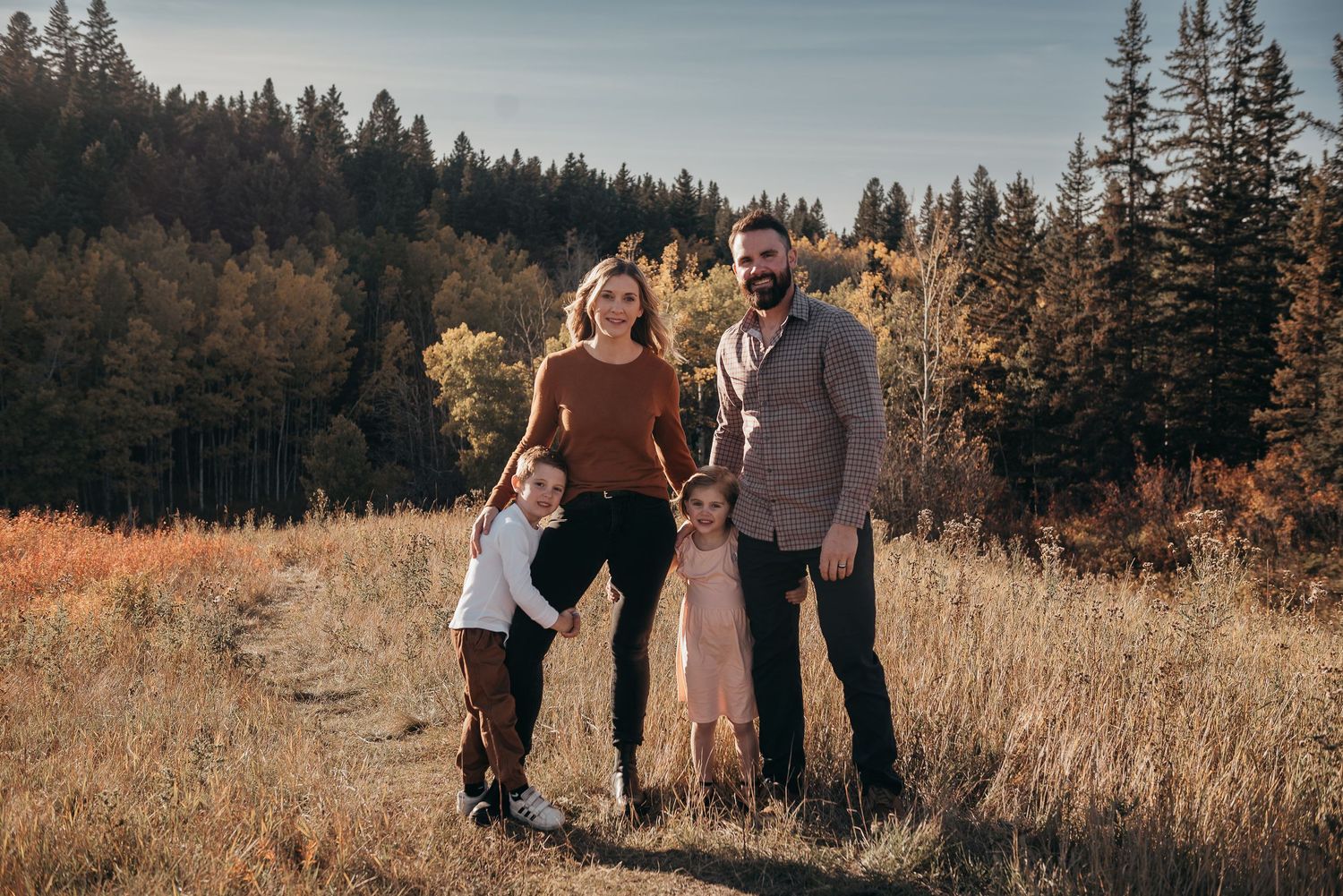 Family of four poses in Calgary autumn meadow with evergreen forest backdrop during golden hour outdoor photo session.