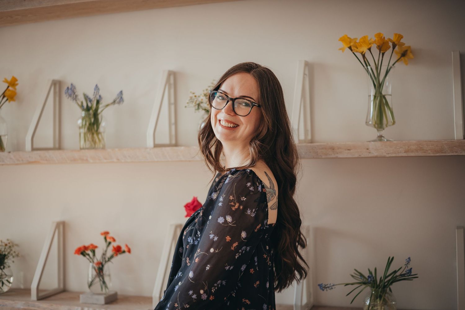 A smiling woman with glasses in a floral shirt poses in a bright room with flower arrangements on wooden shelves.