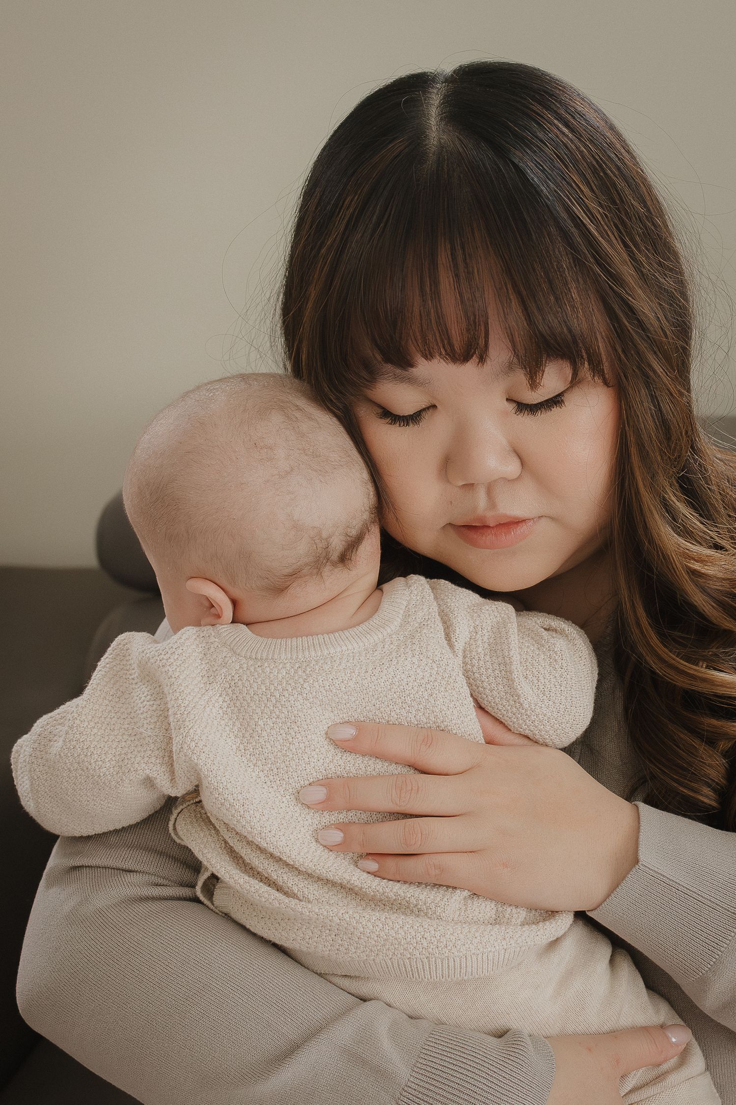 Mother with dark hair tenderly holding and cuddling her newborn baby dressed in white for a lifestyle family photography session in Calgary
