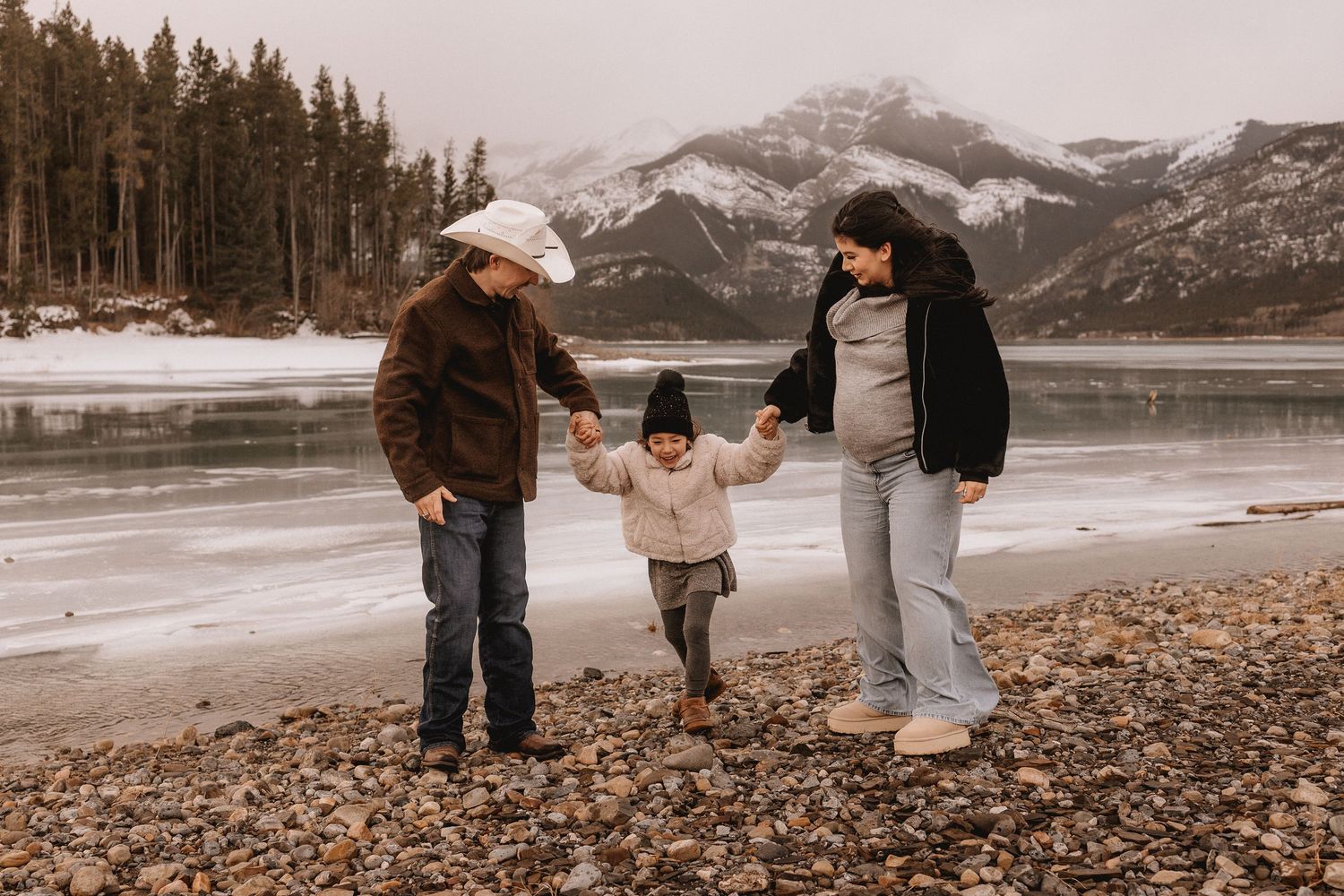 Family of three holding hands on rocky lakeshore with snowy mountains and pine trees in winter backdrop.