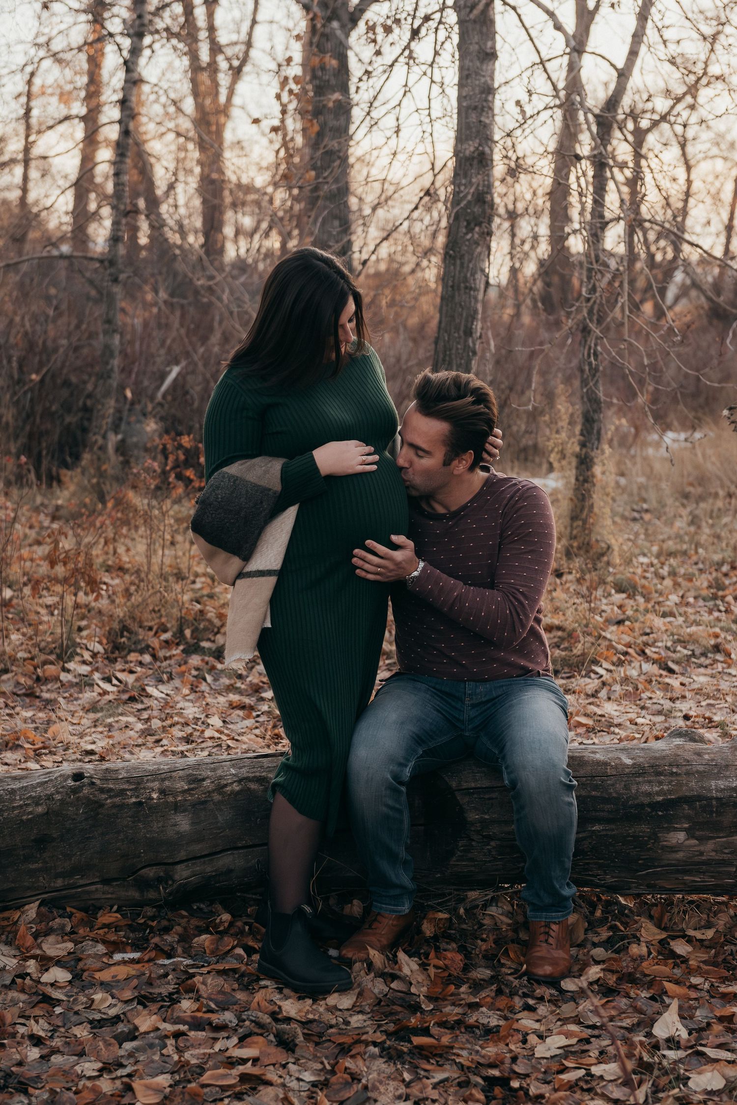 A couple shares an intimate moment while sitting on a fallen log surrounded by autumn leaves.