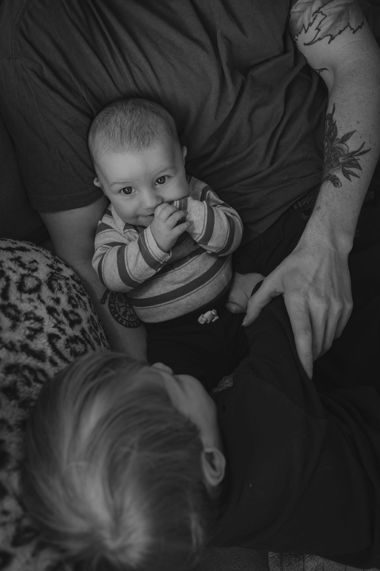 Lifestyle Calgary photography black and white photo of a baby in striped onesie being held by tattooed dad while older brother looks on.