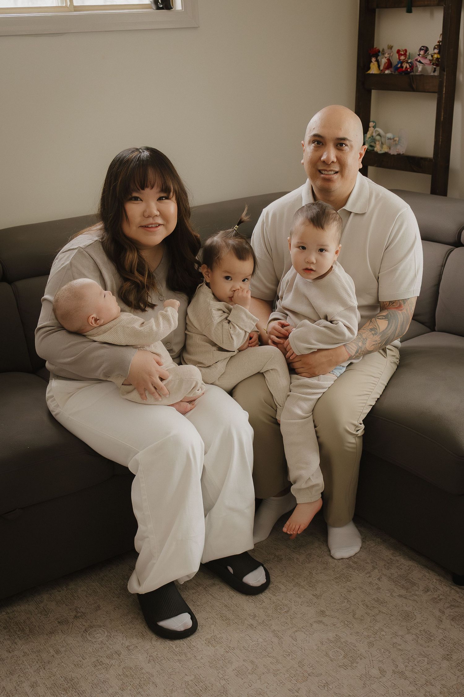 A smiling family of five sits on a gray sofa, parents holding a newborn and two kids dressed in matching neutral outfits.