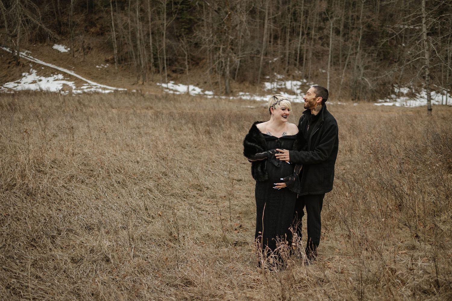 Couple in black outfits embracing in a winter field with snow-dusted hills, maternity photo shoot outdoors.