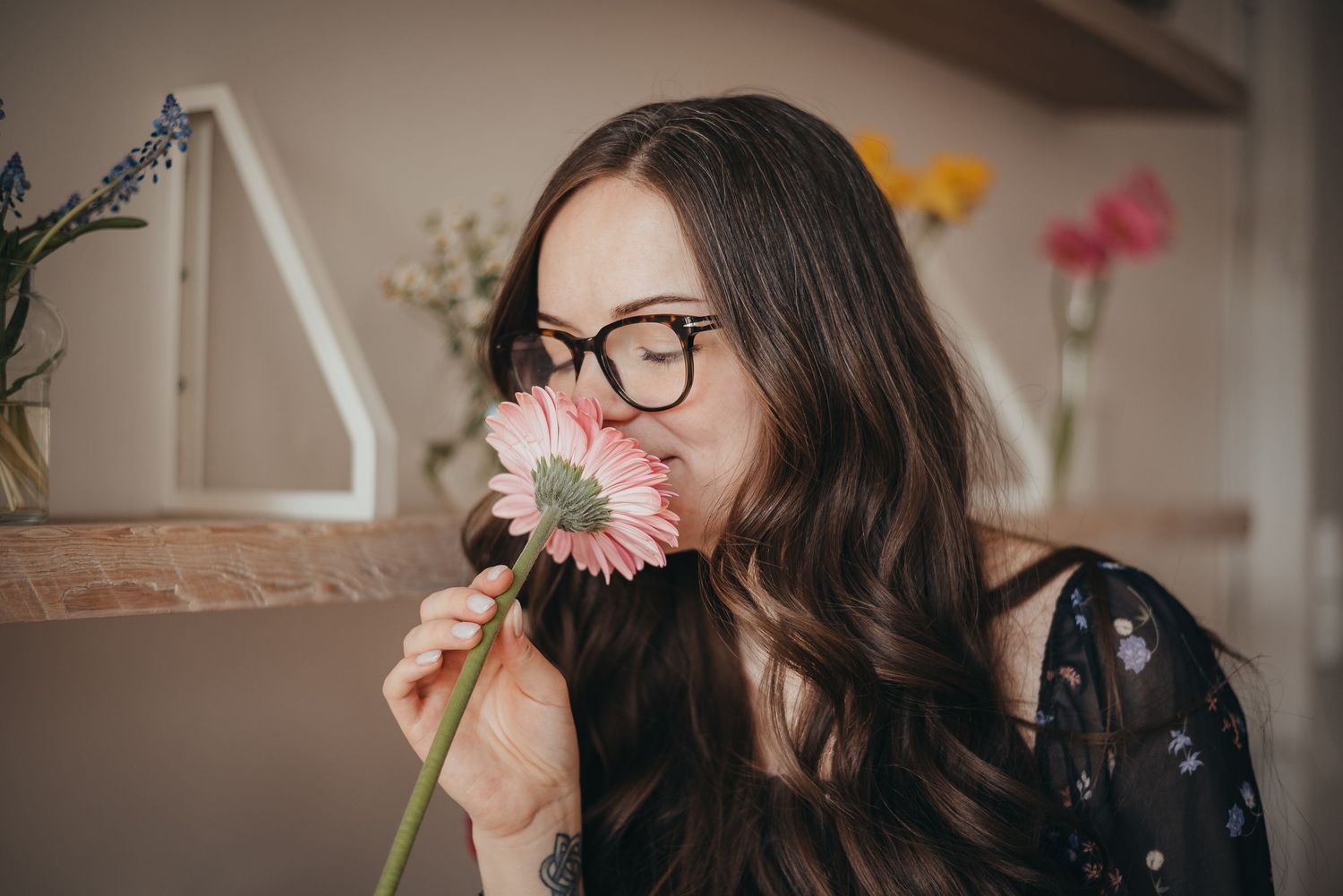 Woman with glasses smelling a pink gerbera daisy in a Calgary studio with colorful flowers in the background.