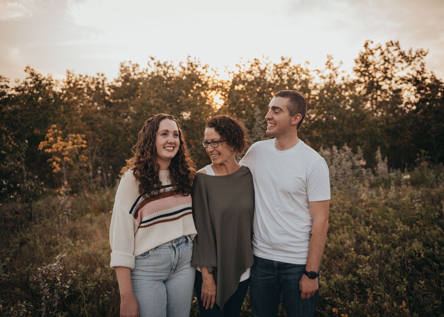 Three people smiling outdoors at golden hour, surrounded by lush greenery and warm sunset light for family portraits.